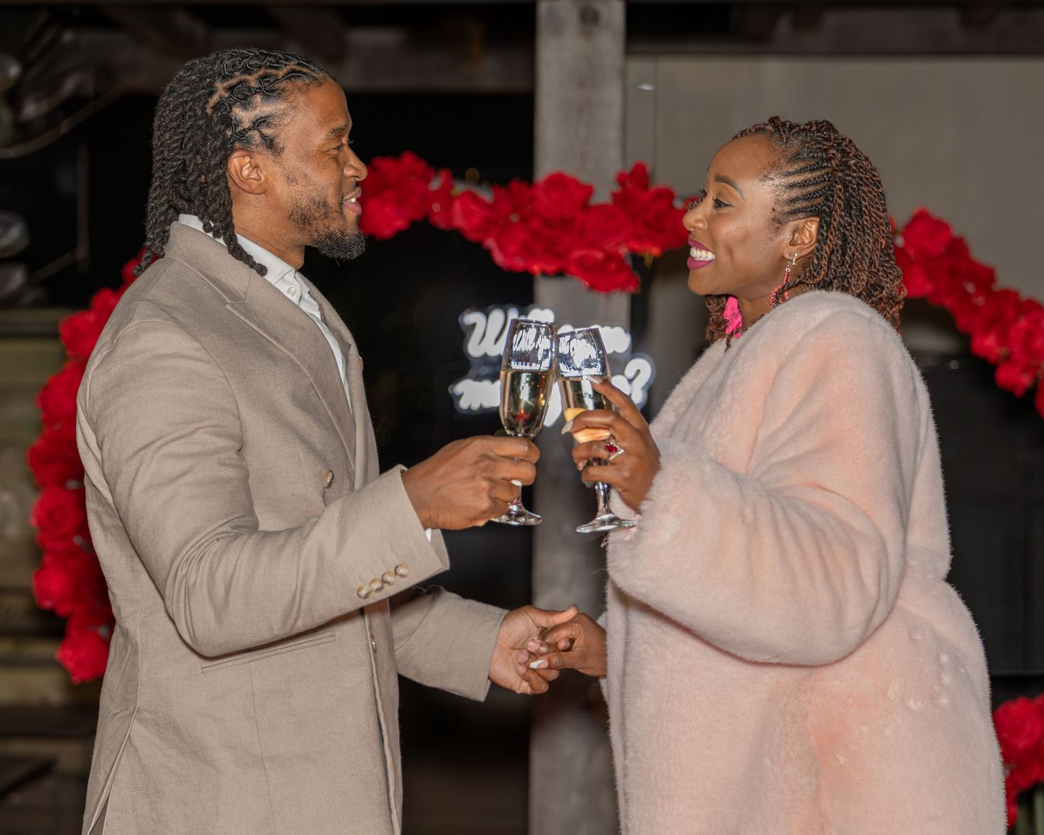 Smiling couple celebrating with champagne glasses at a romantic red rose decorated venue.