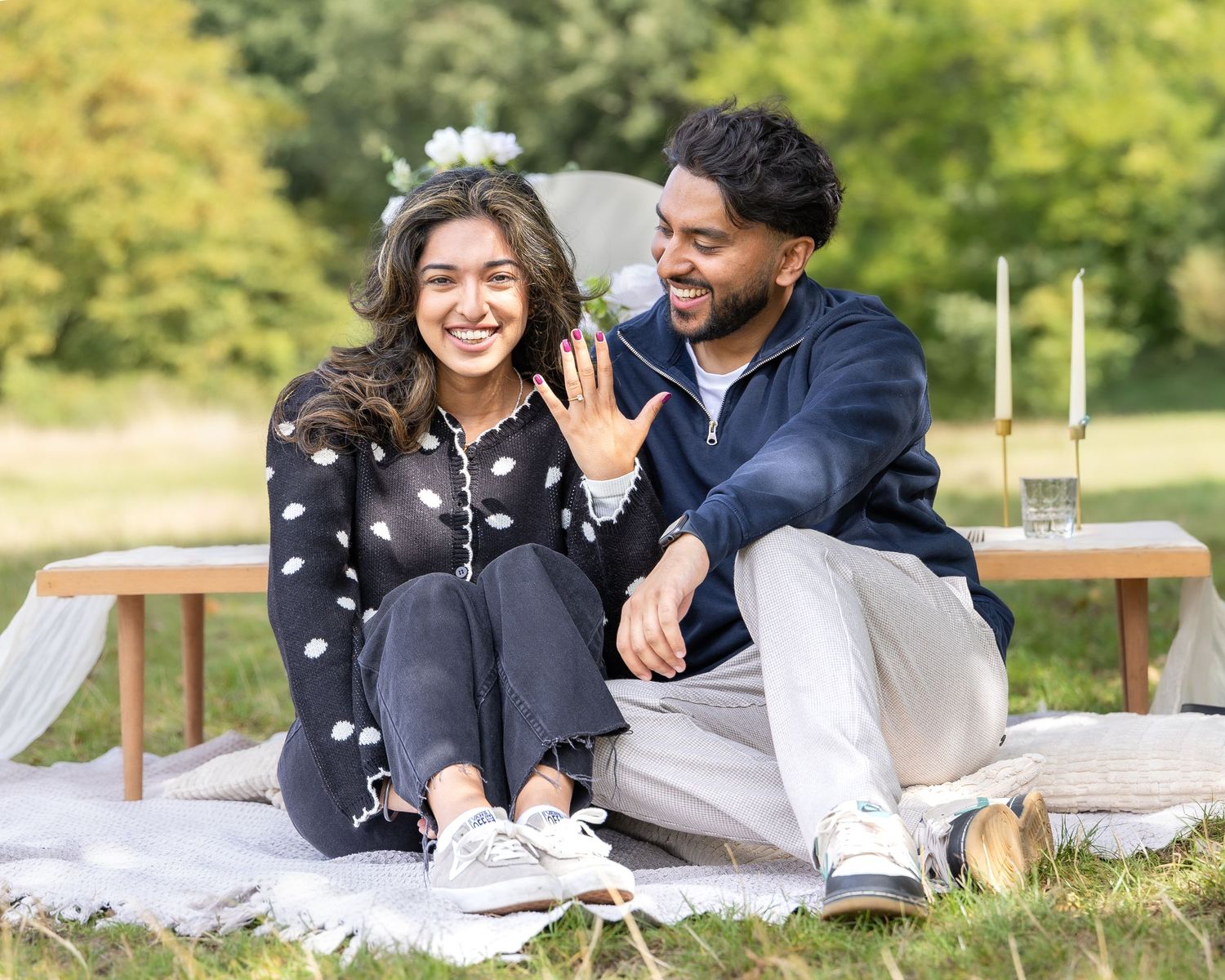 Happy couple showing engagement ring at outdoor picnic proposal, woman wearing floral hair accessory.