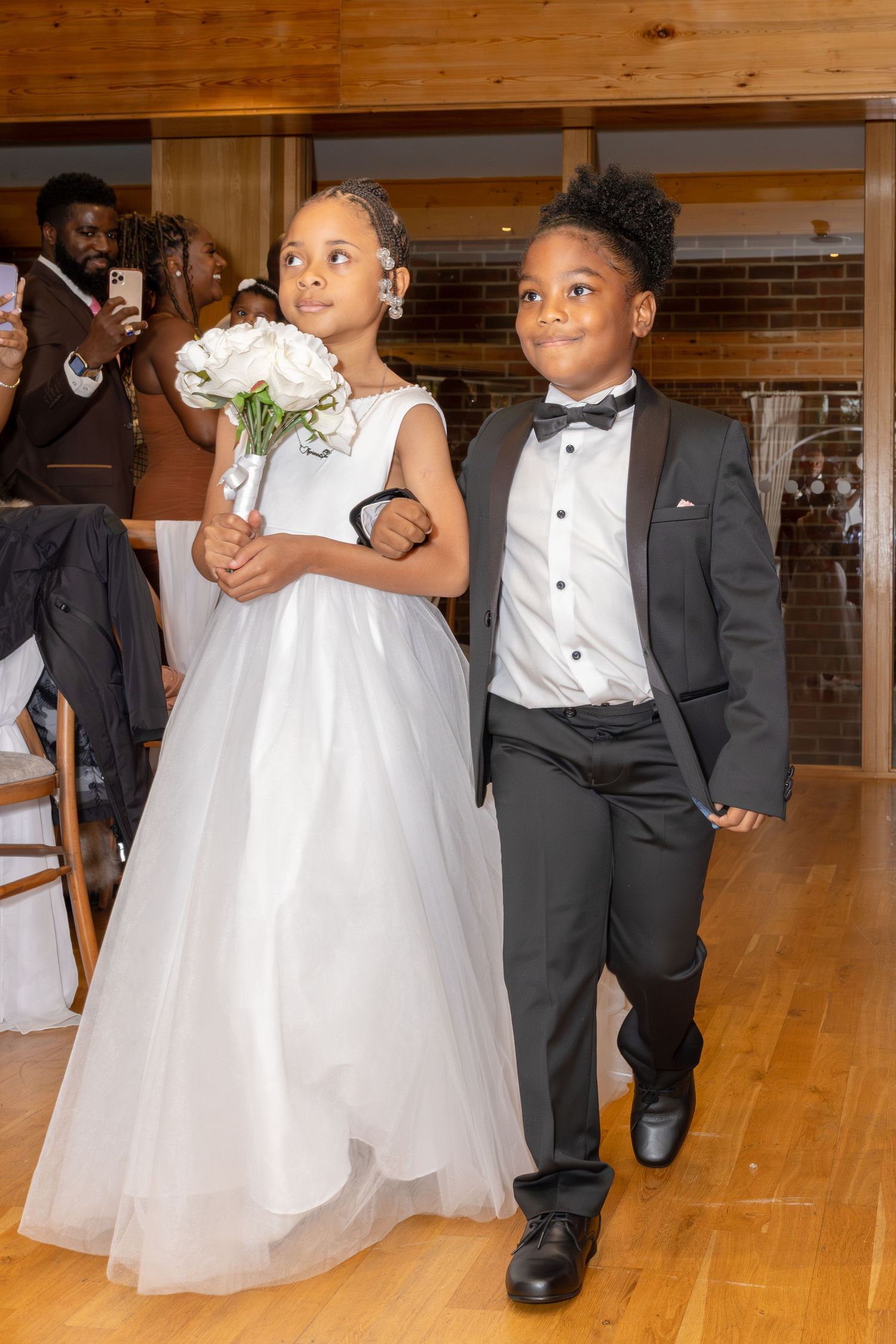 A young flower girl in a white tulle dress and ring bearer in a tuxedo walk down the aisle at a wedding ceremony.