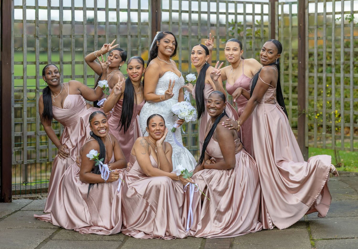 Bride surrounded by bridesmaids in dusty rose satin gowns posing playfully together at an outdoor wedding venue.