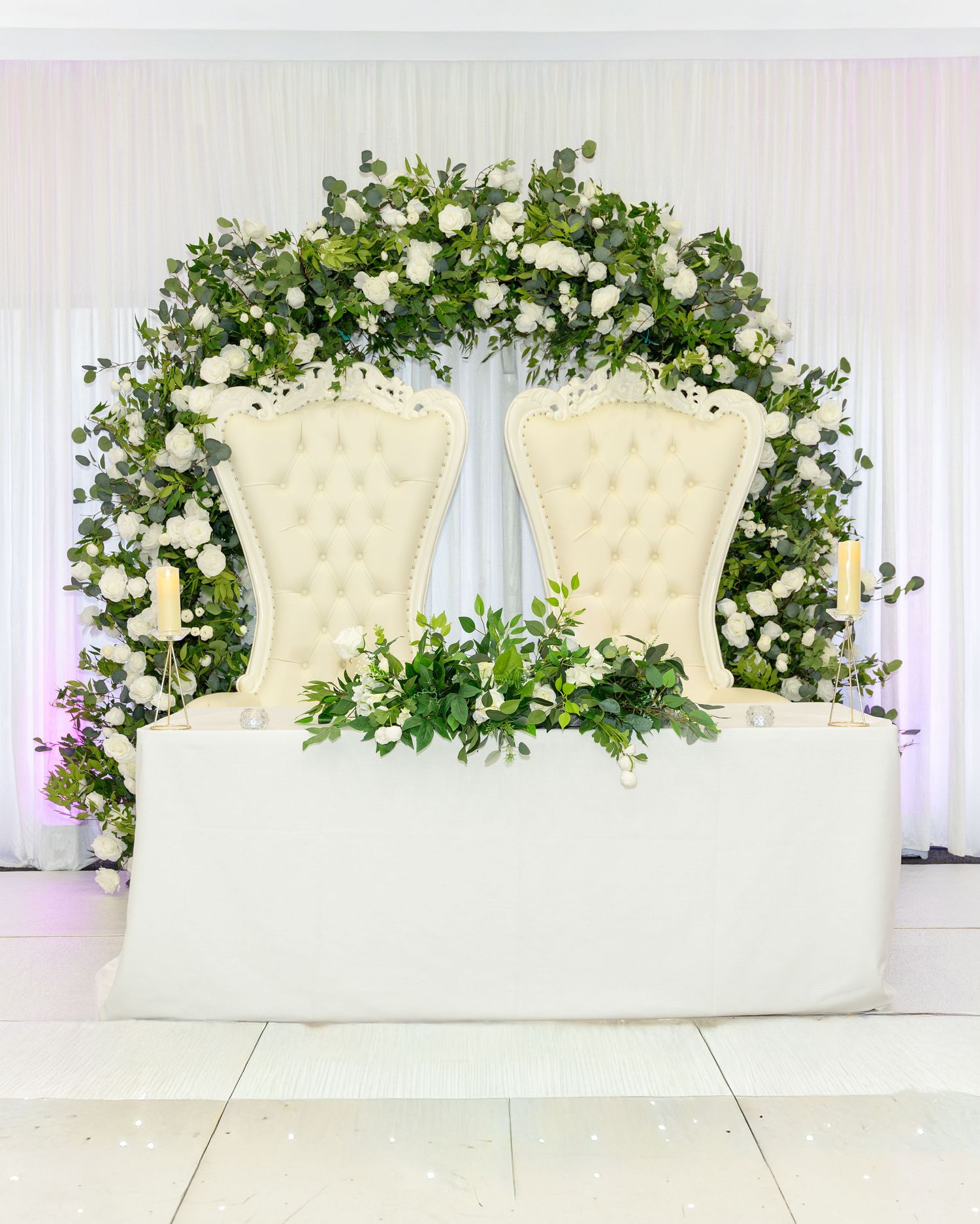 Elegant wedding sweetheart table with white tufted chairs, floral arch of white roses and greenery.