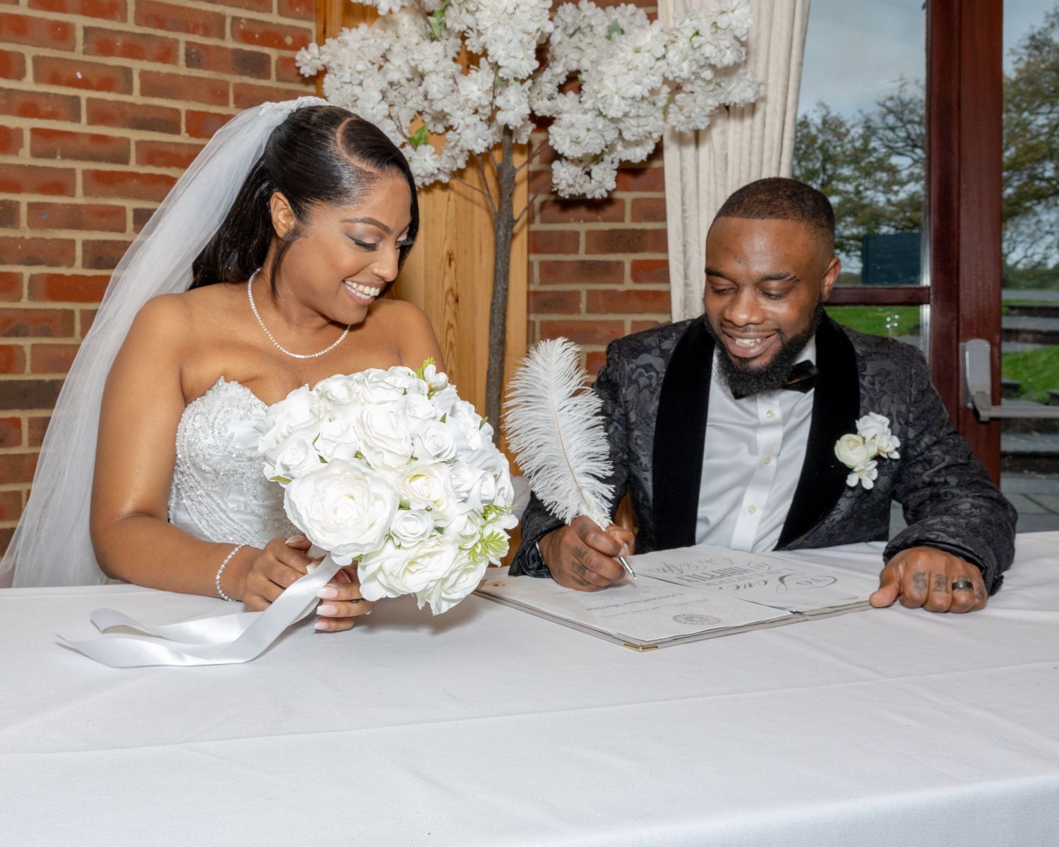 Wedding couple signing marriage documents at table with white floral arrangements.