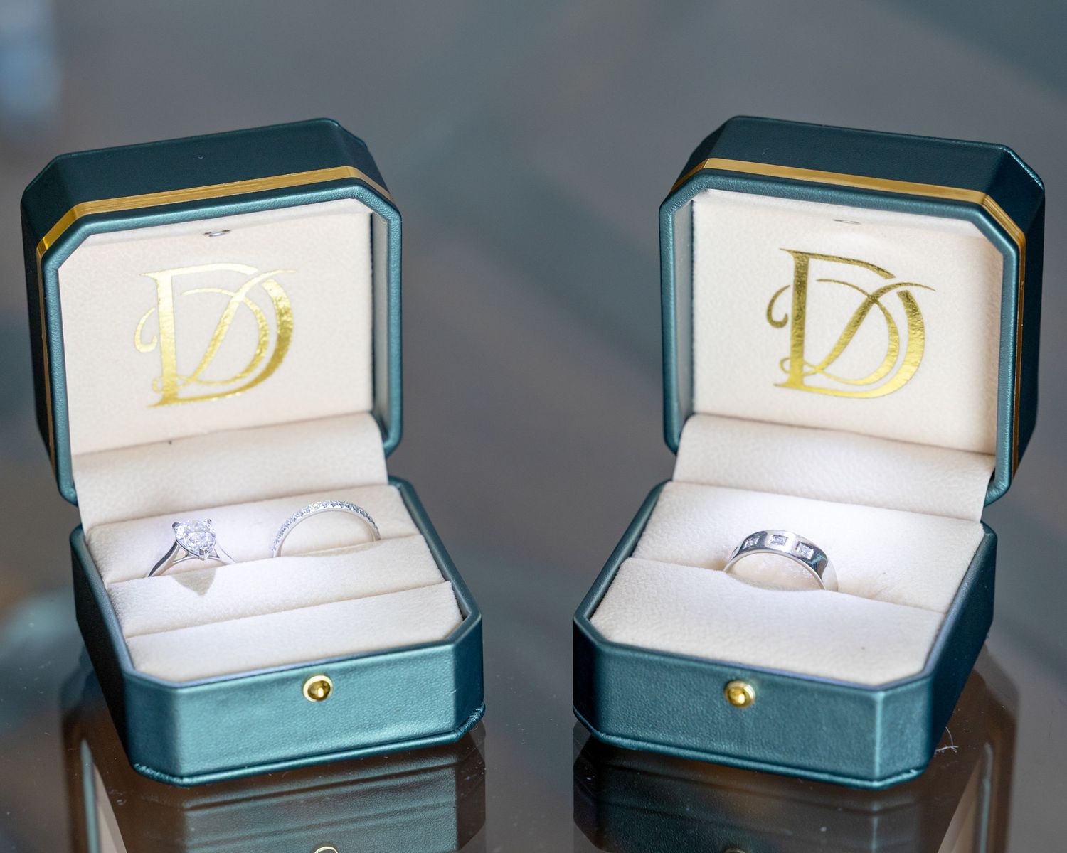 Two elegant teal wedding ring boxes with gold D monogram logos, each containing a silver diamond engagement ring and wedding band.