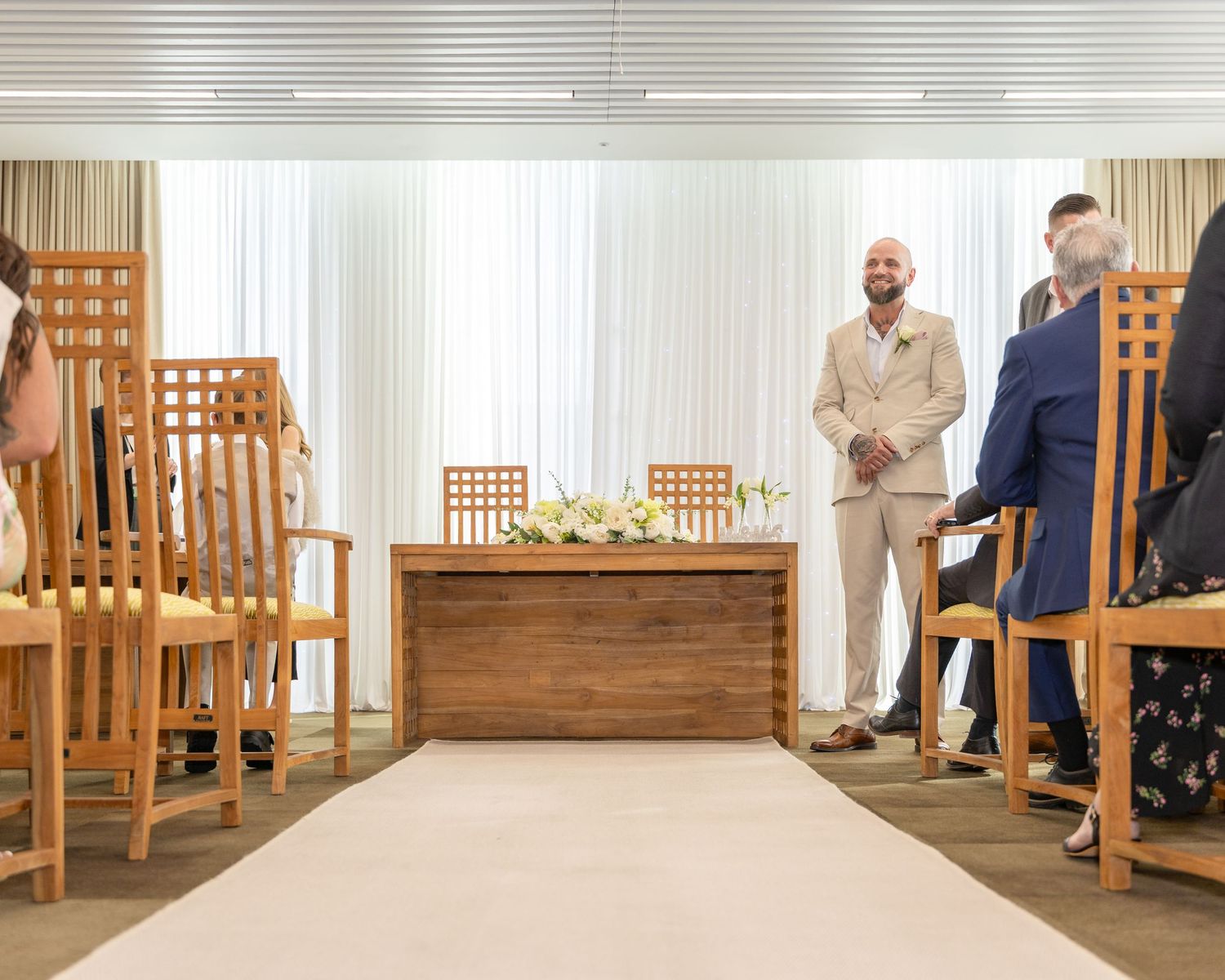 Groom in beige suit smiling at guests during an intimate indoor wedding ceremony.