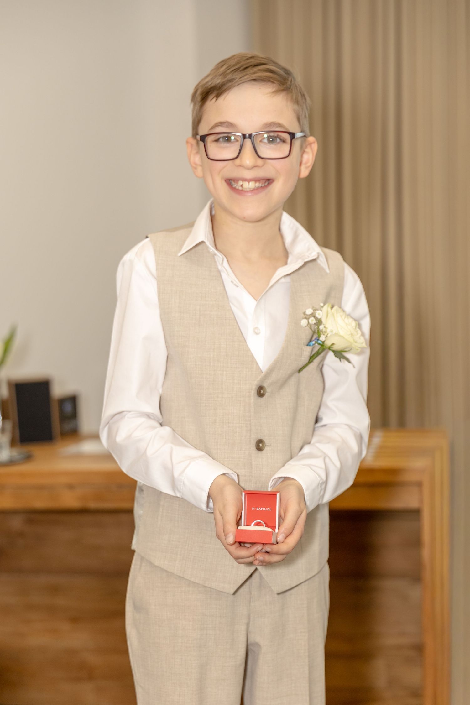 Smiling young boy in a beige vest holding a red ring box at a wedding.