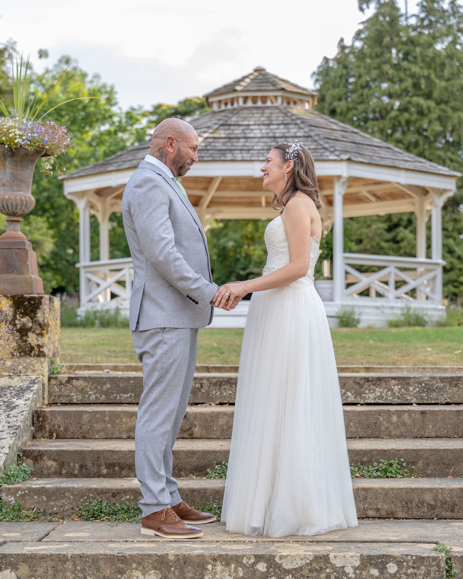 A couple in wedding attire holds hands on stone steps before a white gazebo in a lush garden.