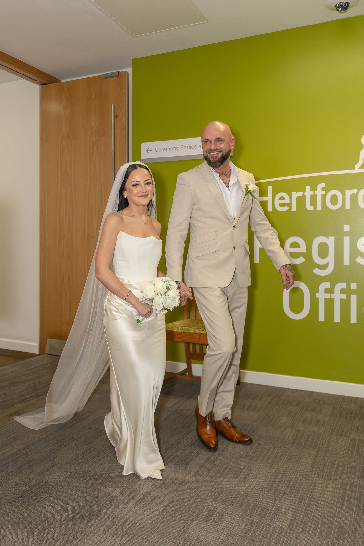 Bride and groom pose smiling at Hertfordshire Register Office entrance after their wedding ceremony.