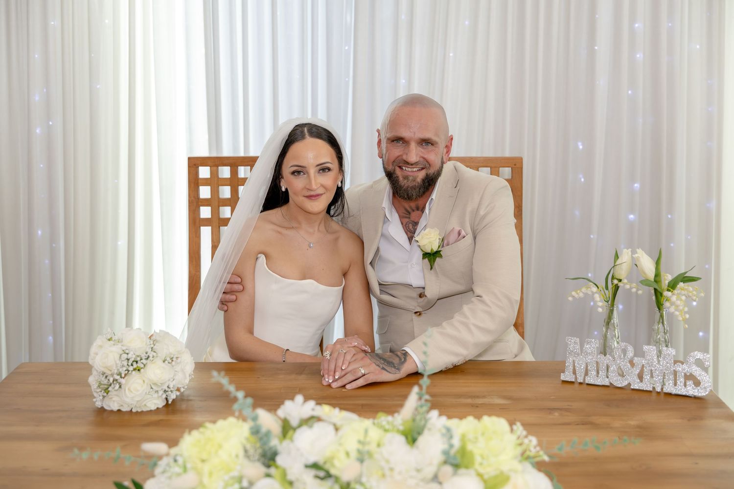 Newlyweds pose at table with Mr and Mrs sign and white bouquet after ceremony.
