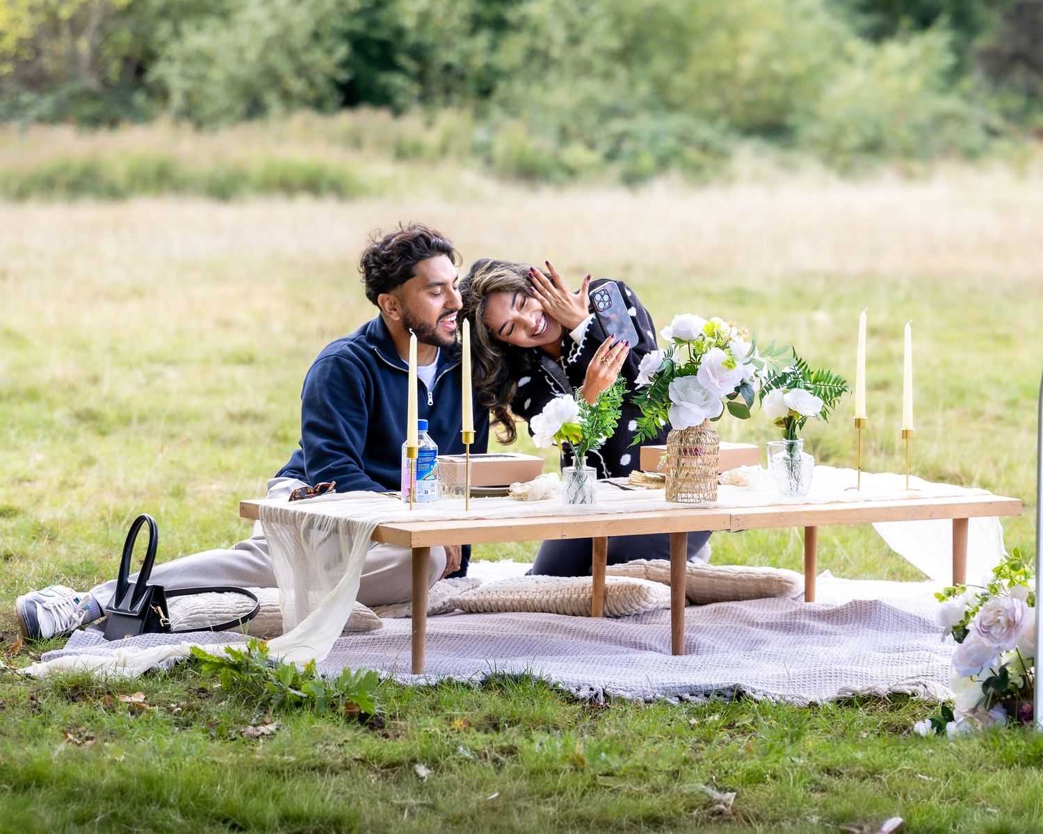 Romantic outdoor picnic proposal setup with flowers, candles, and couple embracing on white blanket.