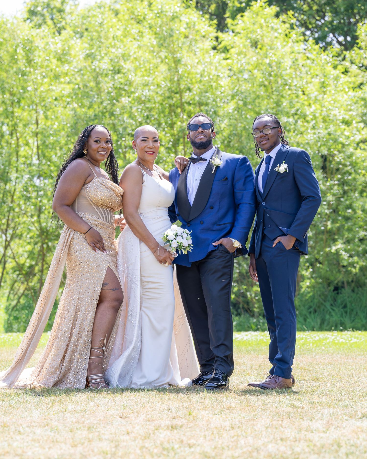 Wedding party poses outdoors in formal attire, bride in white gown with groom and guests in navy suits.