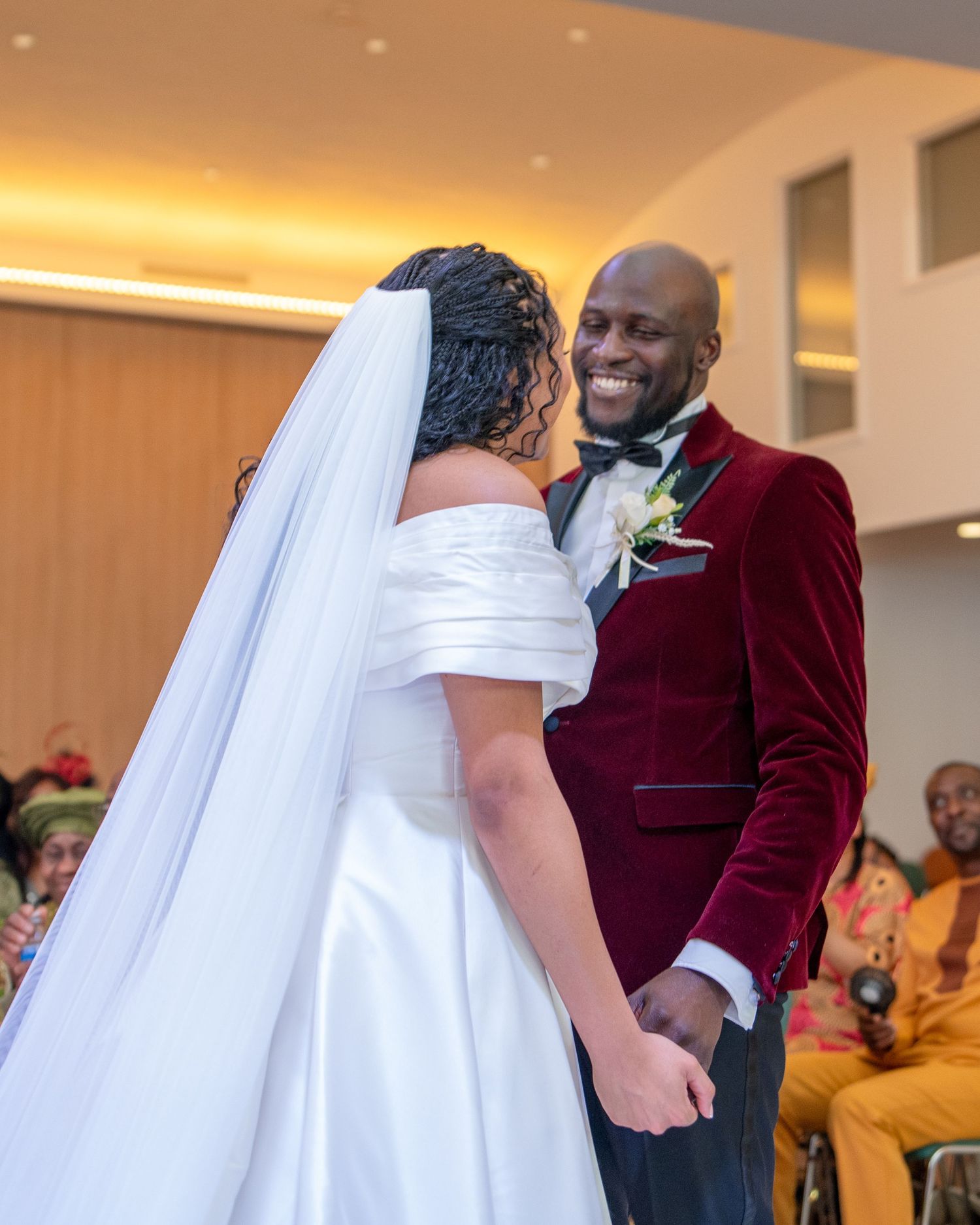 A smiling groom in a burgundy velvet tuxedo holds hands with his bride in a white off-shoulder gown at their wedding.