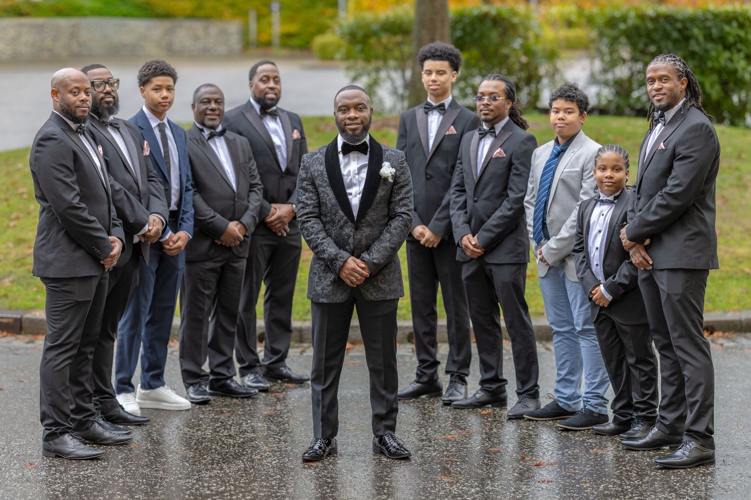 Groom in patterned tuxedo posing with his groomsmen and family outdoors on a wet autumn day.