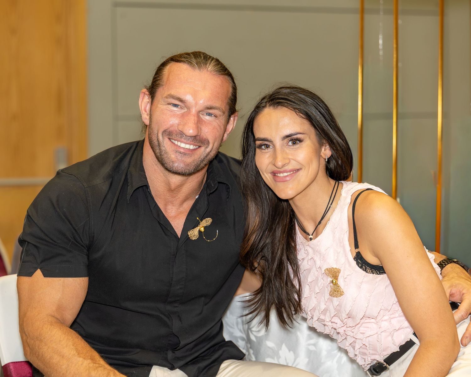Smiling couple poses together at an indoor event, man in black shirt and woman in light pink dress.