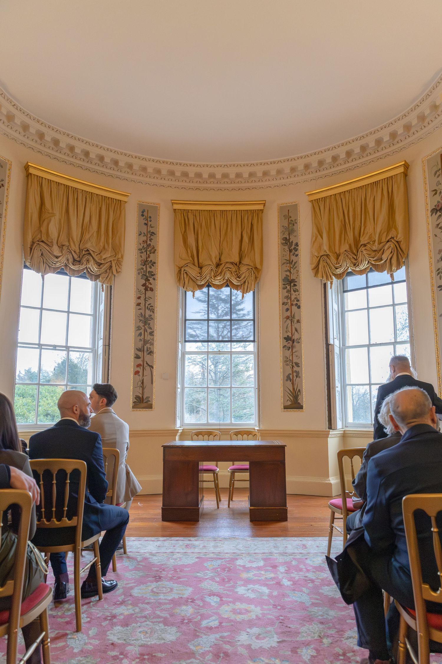 Wedding ceremony in an elegant oval room with tall windows and guests seated in rows.