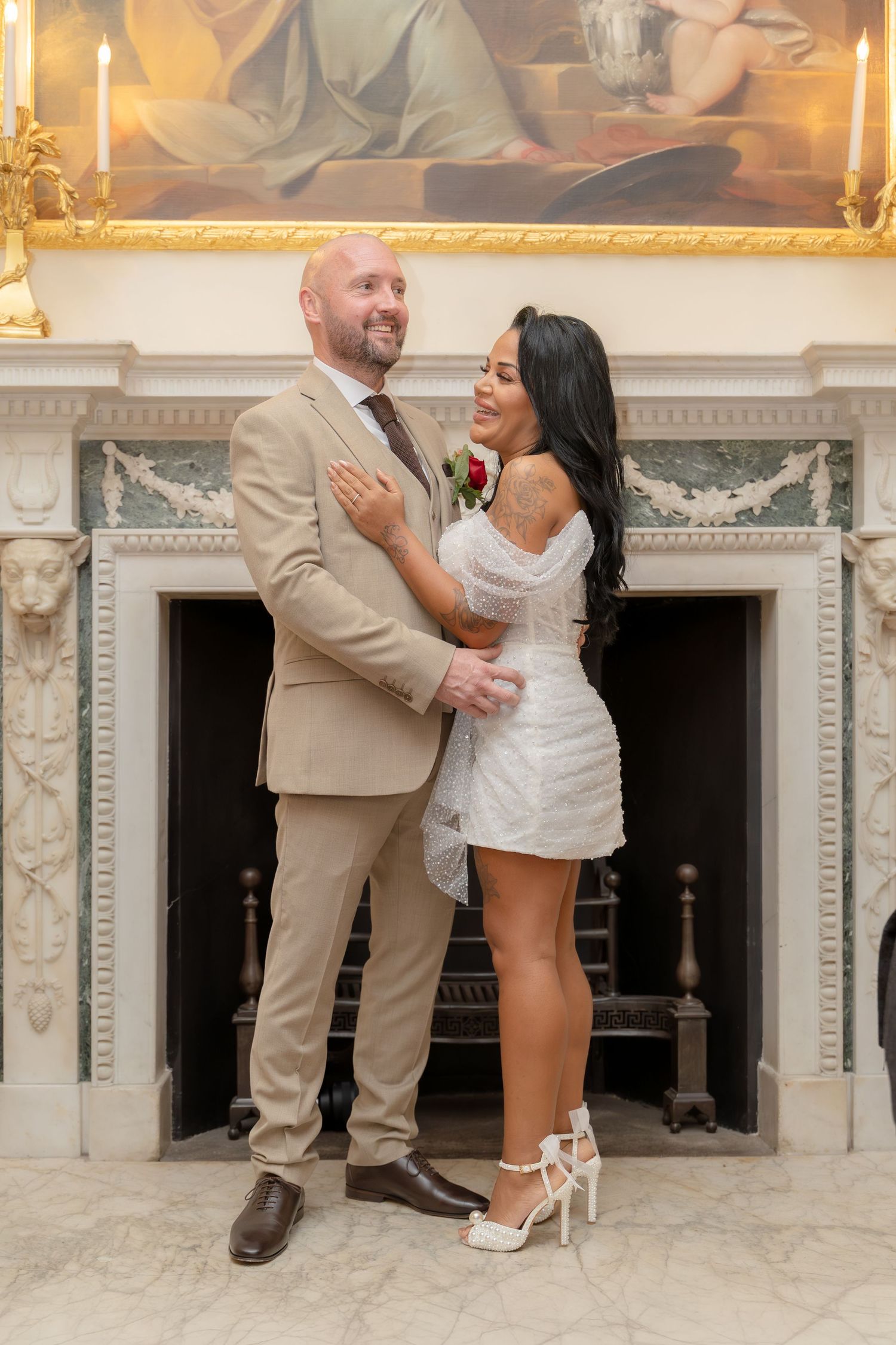 Smiling newlywed couple embracing before decorative fireplace, groom in beige suit, bride in white mini dress.