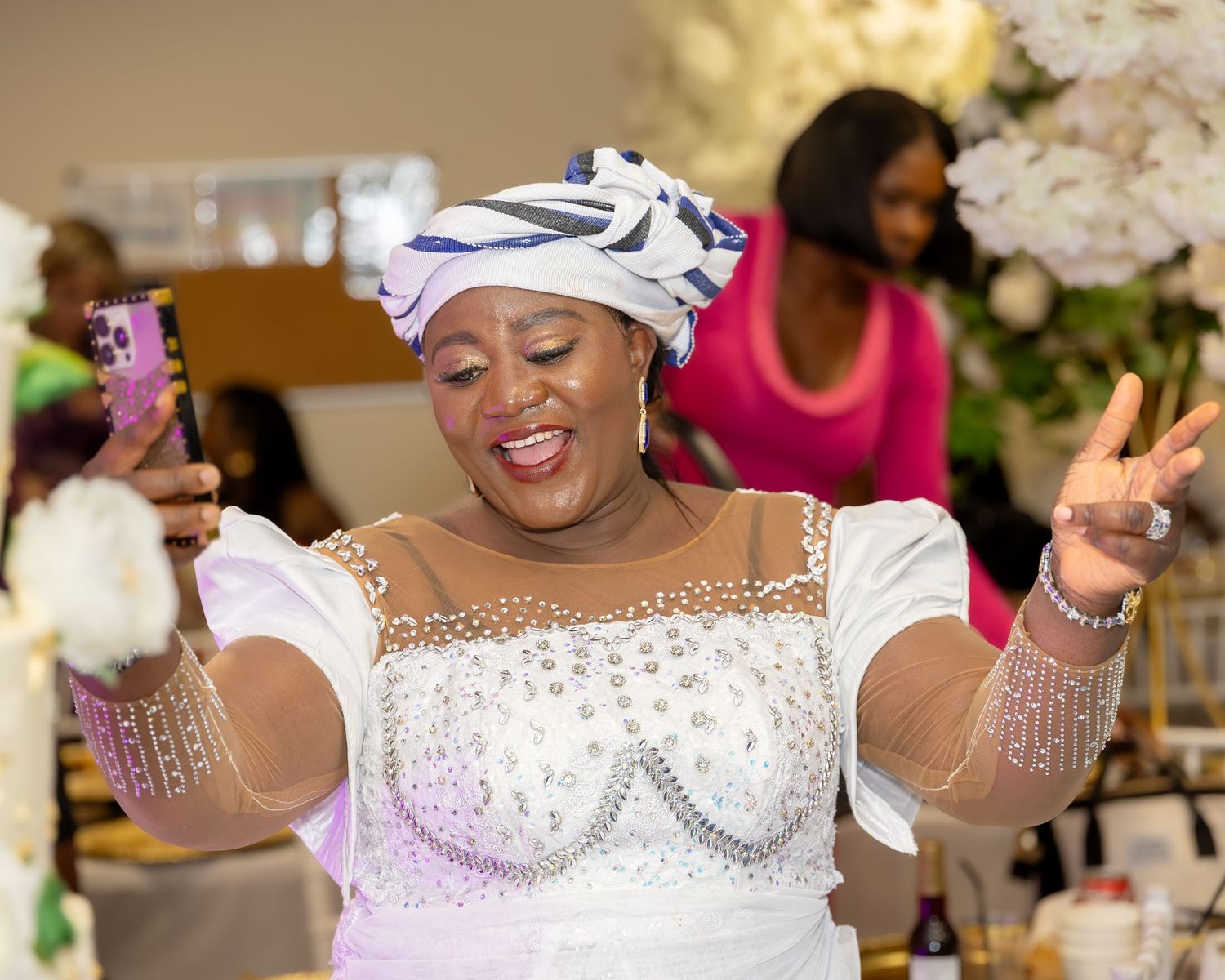 A joyful woman in a white beaded gown and blue head wrap dances while holding a phone at a Nigerian celebration.