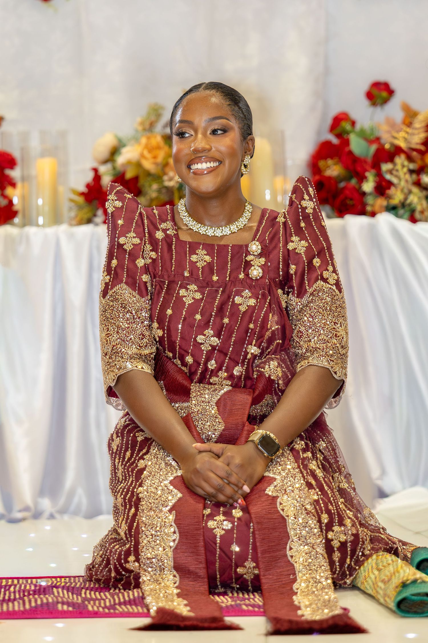 Smiling bride in ornate burgundy and gold African traditional wedding dress kneels before a floral altar.