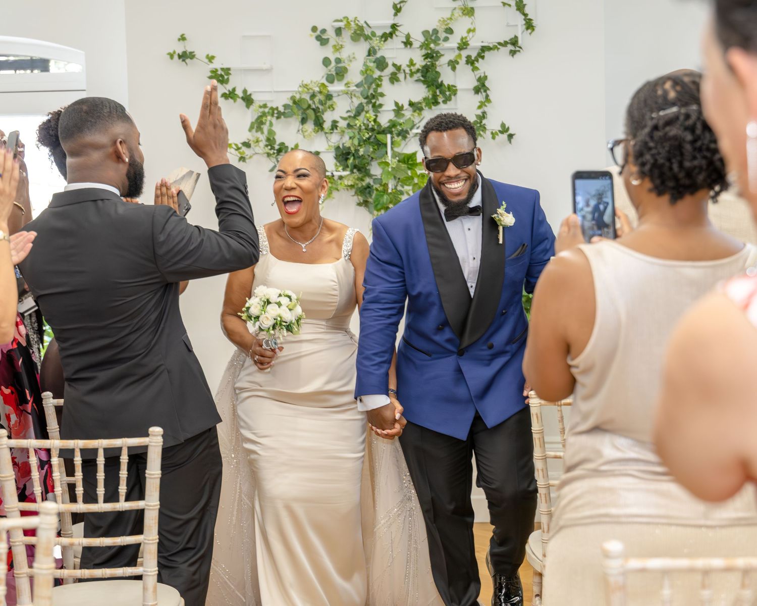 Joyful newlyweds walk down the aisle, bride in white gown holding bouquet, groom in blue suit, guests celebrating.