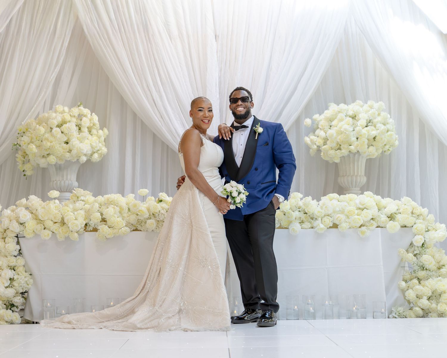 Elegant wedding couple posing before white floral arrangements and draped backdrop in formal attire.