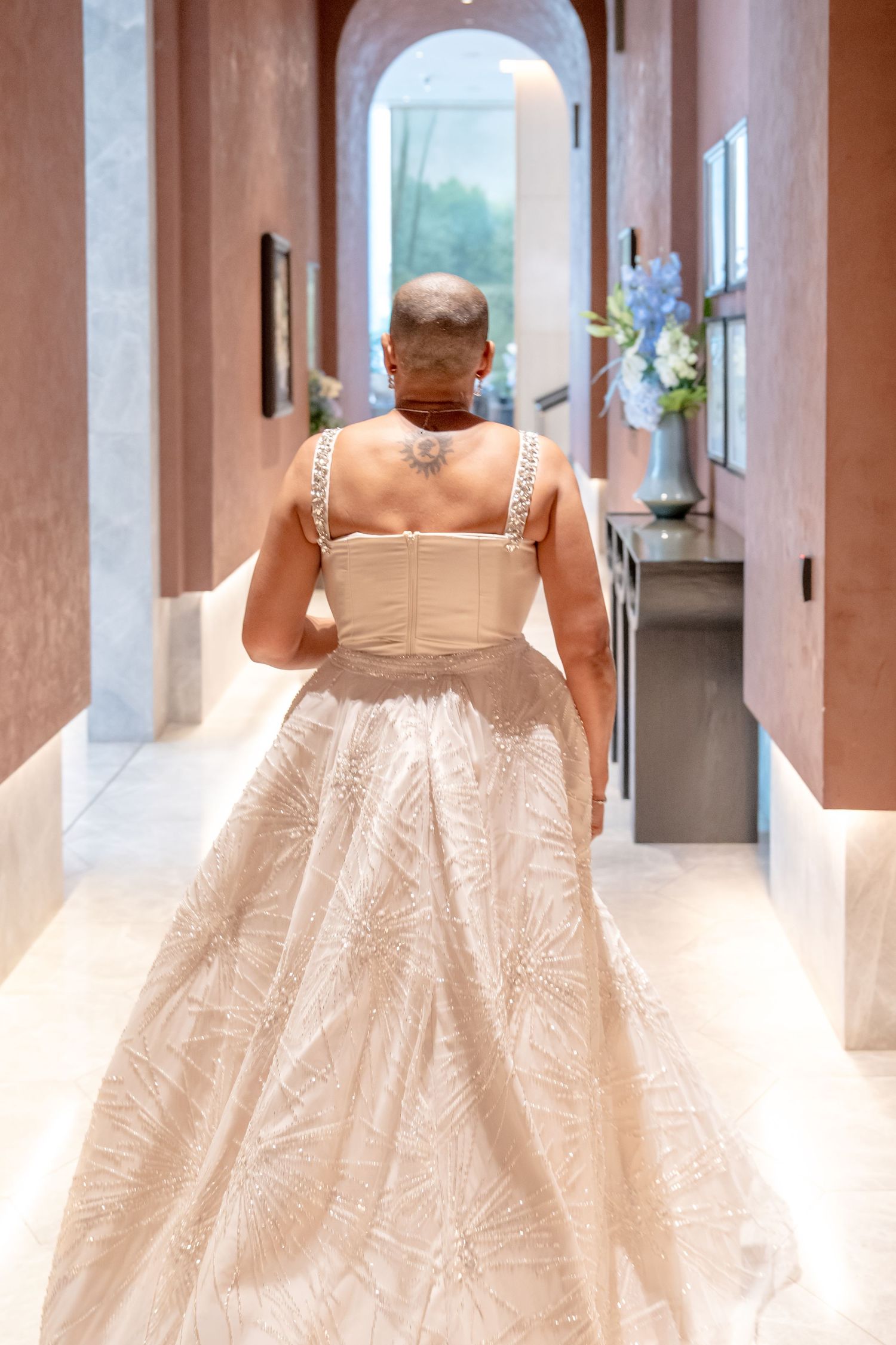Person in elegant champagne ballgown with floral embroidery and jeweled straps walking through luxurious hotel corridor.