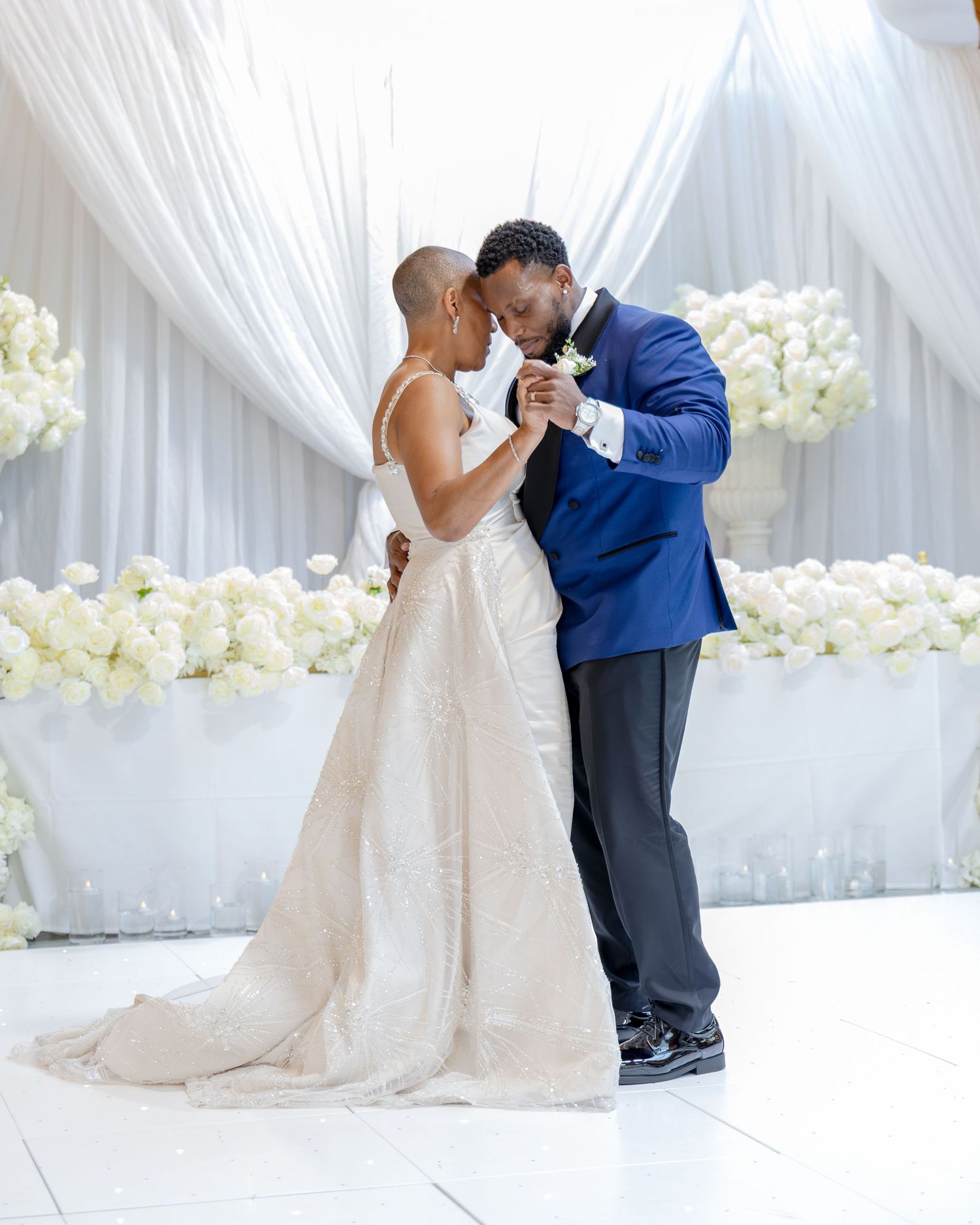 A bride and groom share their first dance surrounded by white floral arrangements and elegant draped fabric backdrop.
