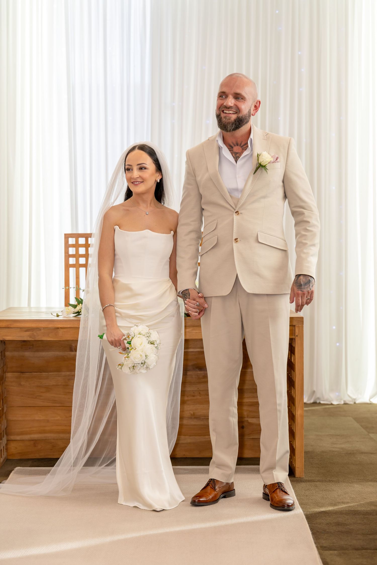 Bride in white gown and groom in beige suit hold hands at wedding altar with white drape backdrop.