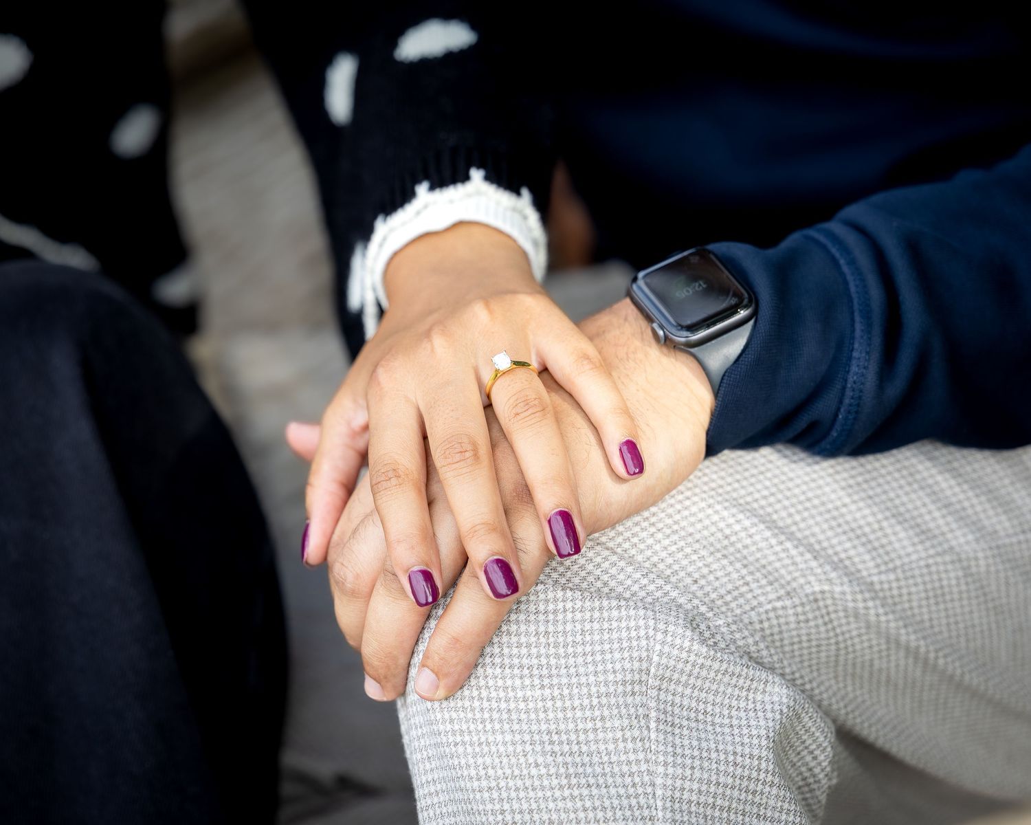 Close-up of couple holding hands showing gold engagement ring with purple nail polish outdoors.