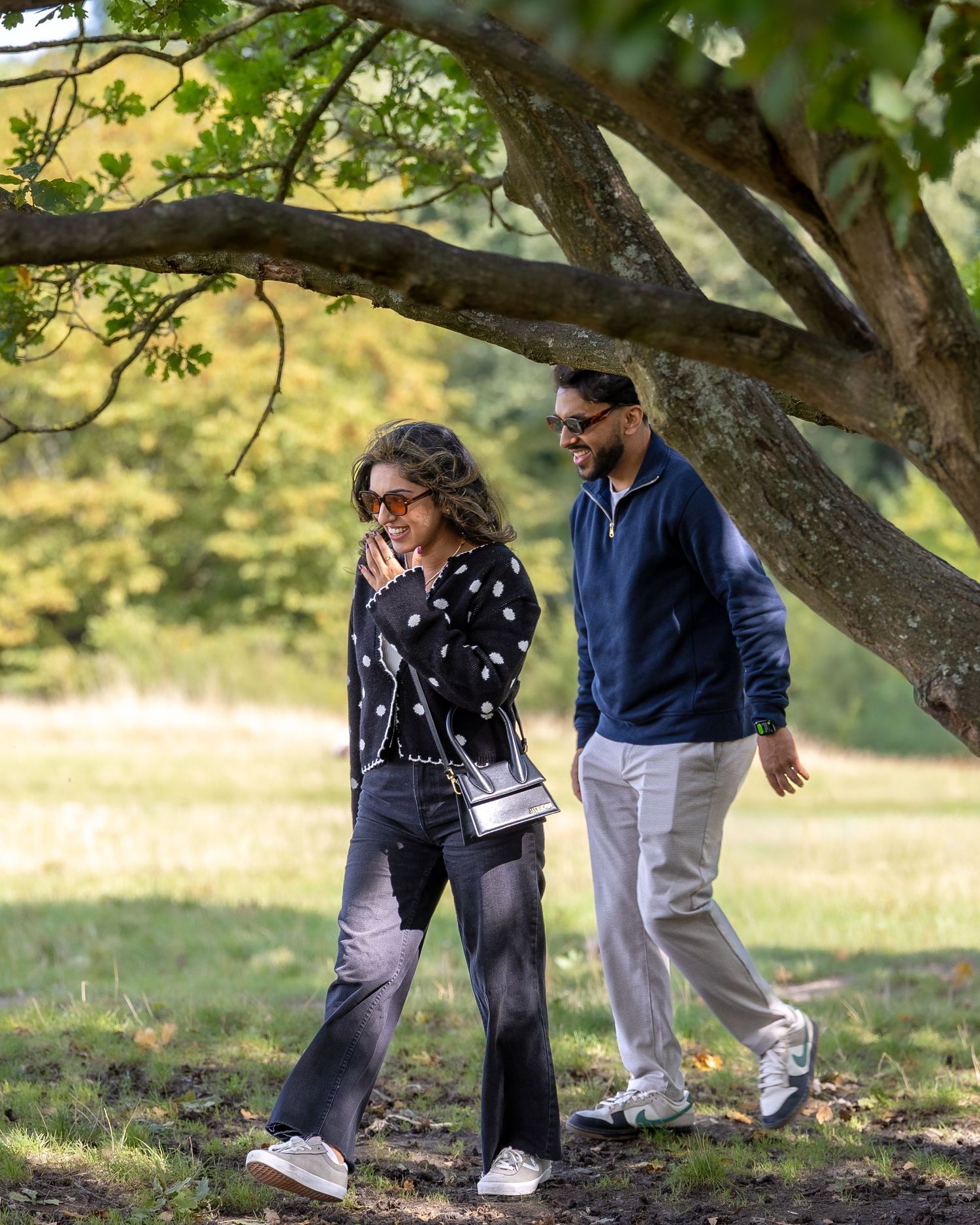 Couple walking together under a large tree in a lush green park on a sunny autumn day.