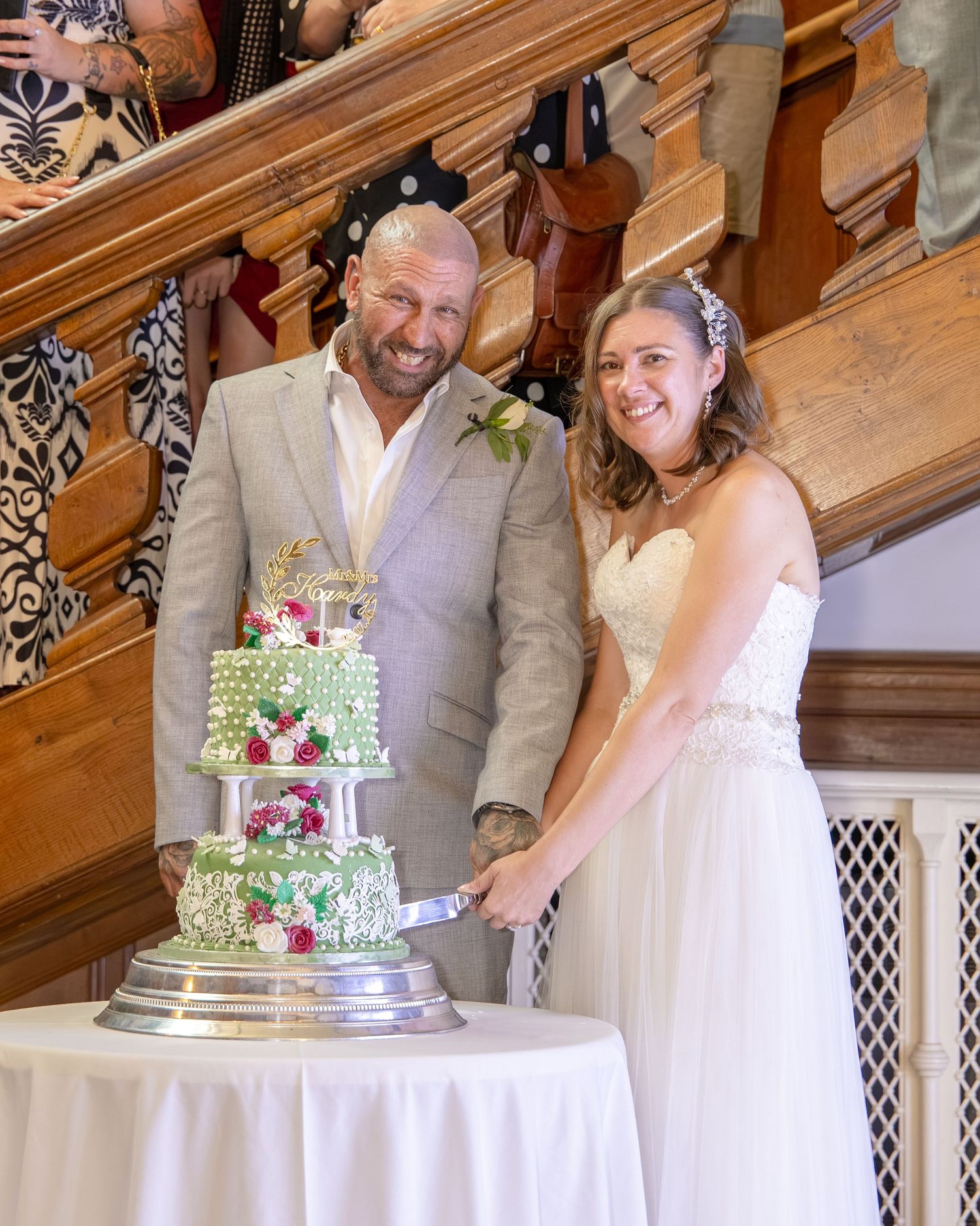 Bride and groom smiling while cutting a decorated three-tier wedding cake near an elegant wooden staircase.