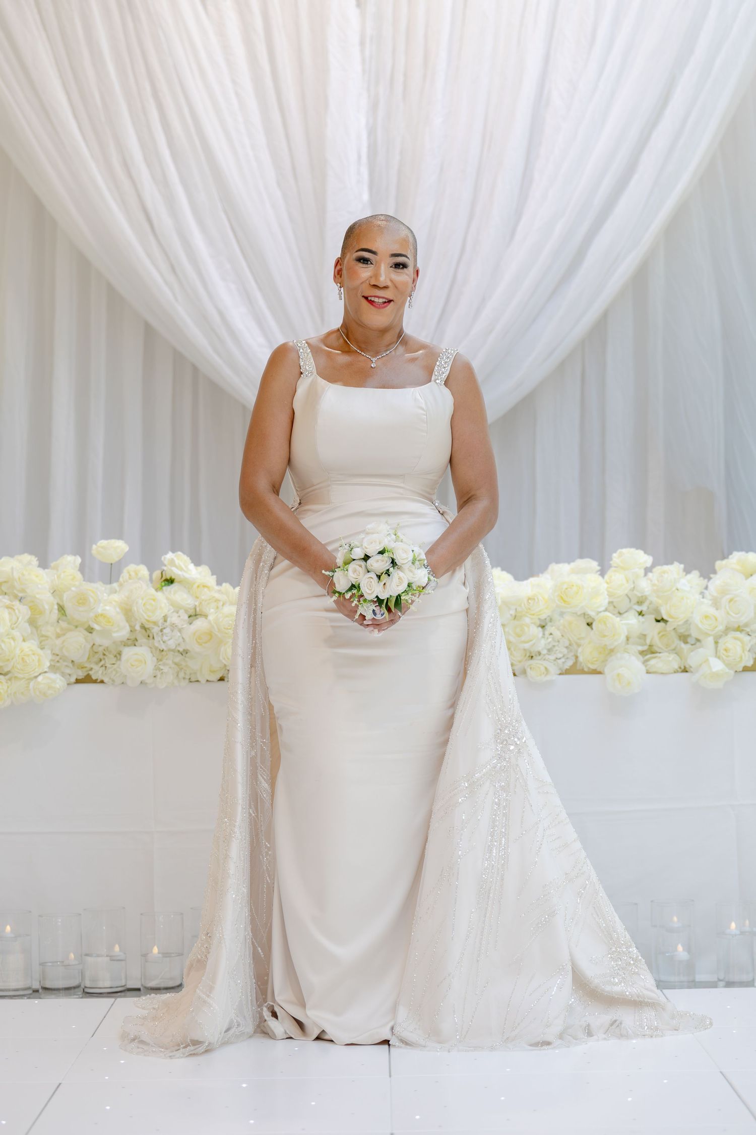 Bride in white sheath gown with lace overlay holds bouquet before floral backdrop at elegant wedding ceremony.