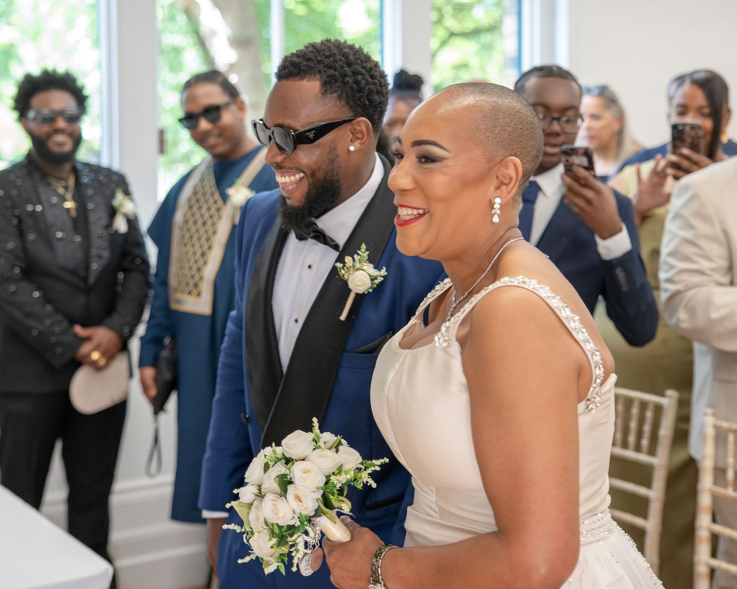 Happy couple smiles during wedding ceremony, groom in blue suit, bride in white gown holding bouquet, guests celebrating behind them.