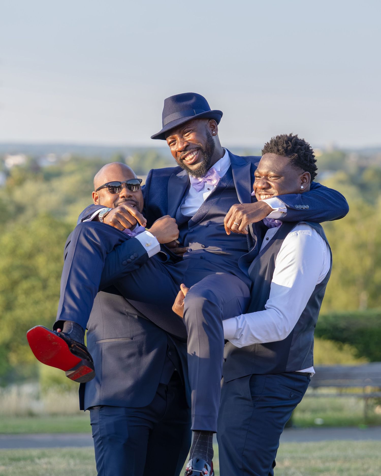 Three well-dressed men in navy suits celebrating outdoors, two lifting the groom in a joyful wedding party moment.