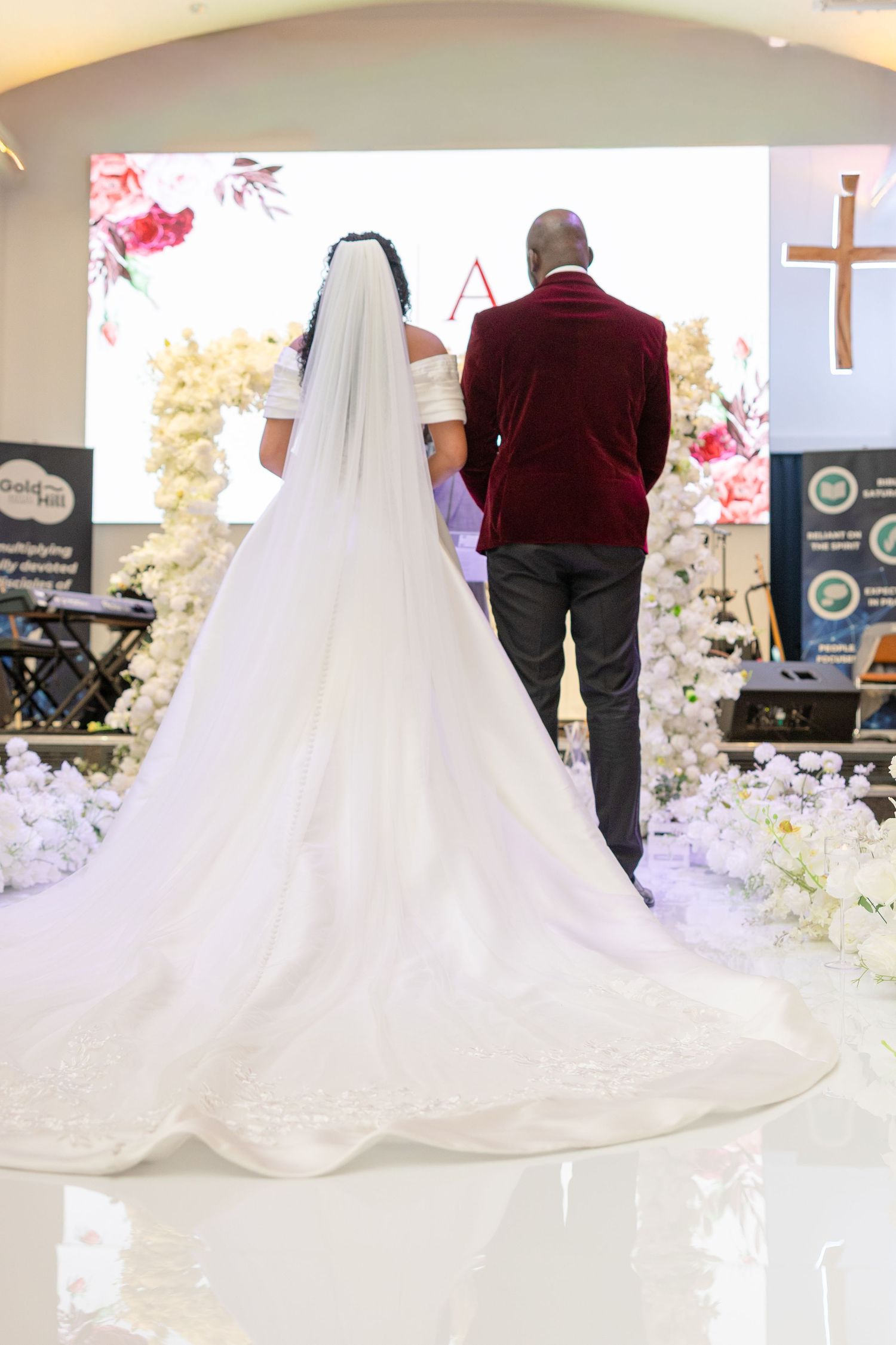 Bride in white gown with long veil stands with groom in burgundy suit at church wedding altar.