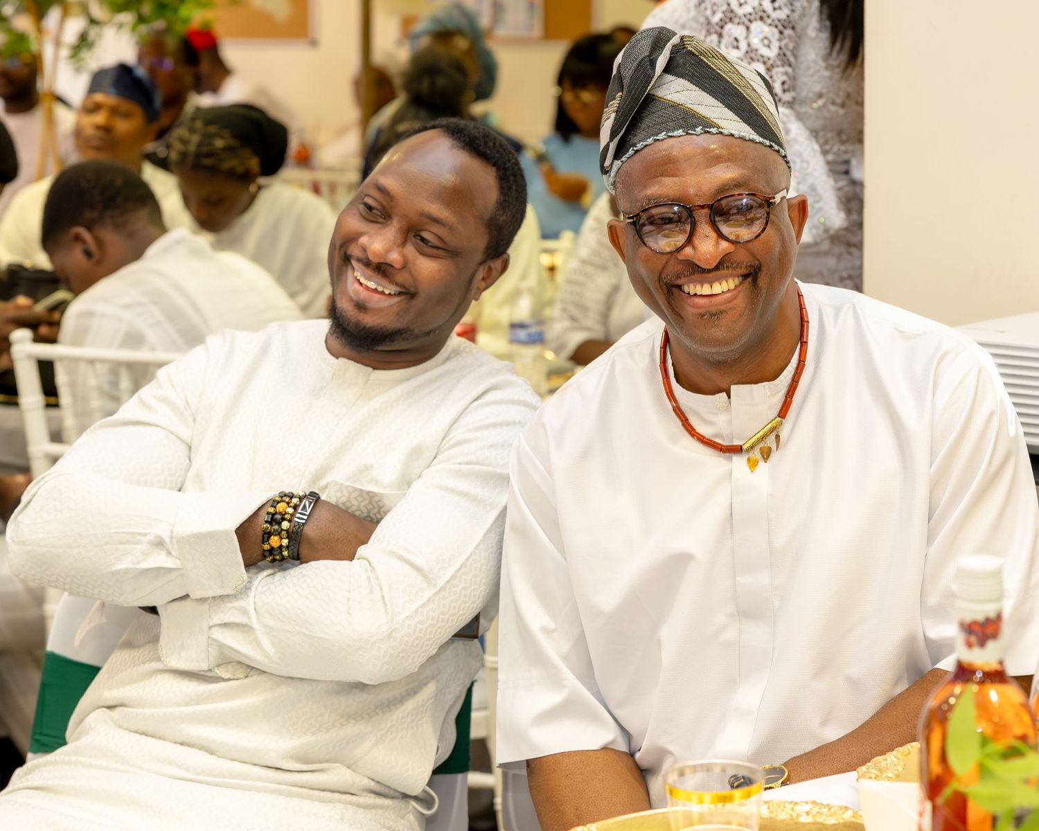 Two Nigerian men in white traditional attire smiling together at a cultural wedding celebration.