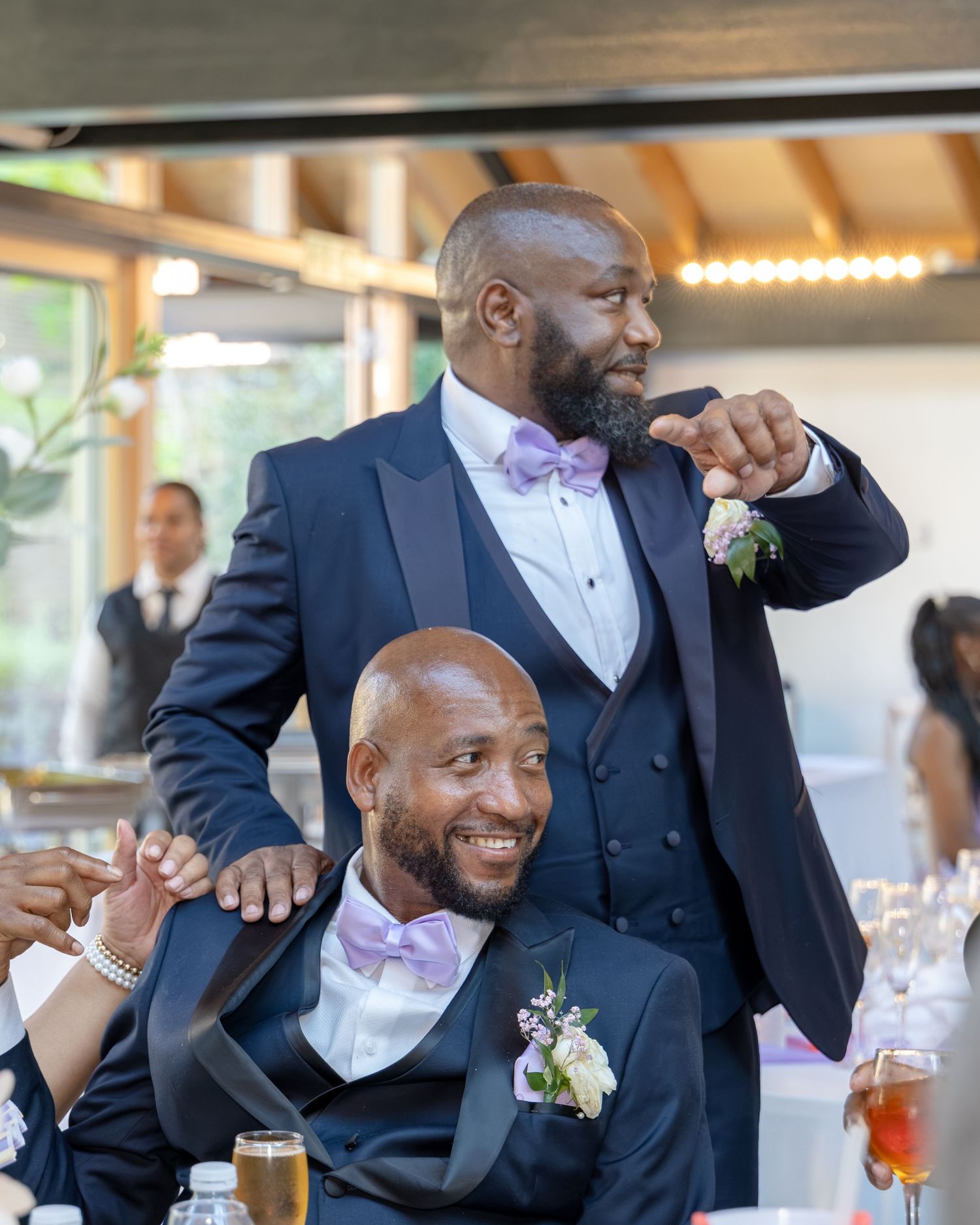 Two men in navy suits with lavender bow ties at a wedding reception, one standing and pointing while the other sits smiling.