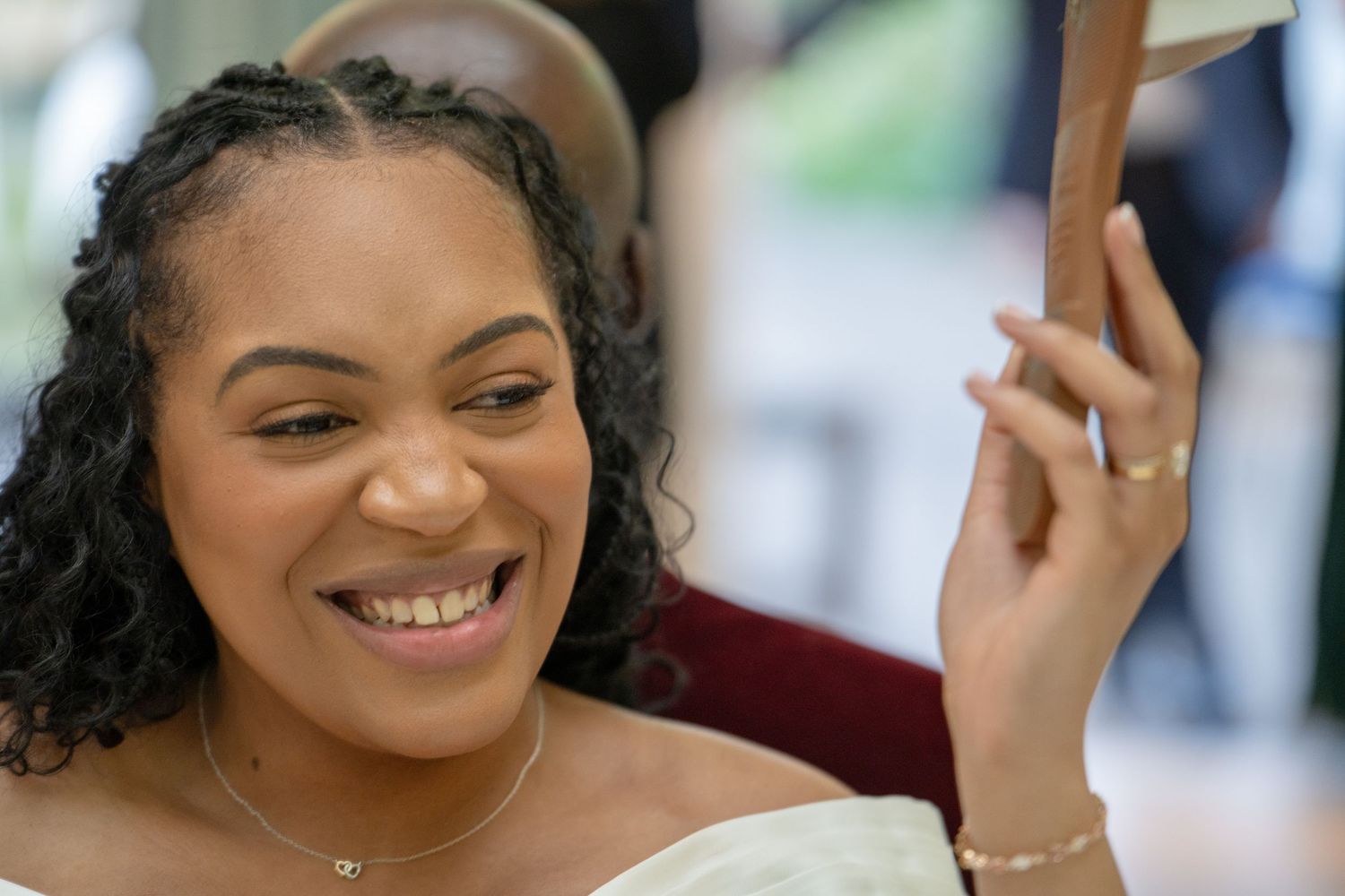 A smiling young woman with natural hair holds a mirror, wearing a white top and delicate gold jewelry.
