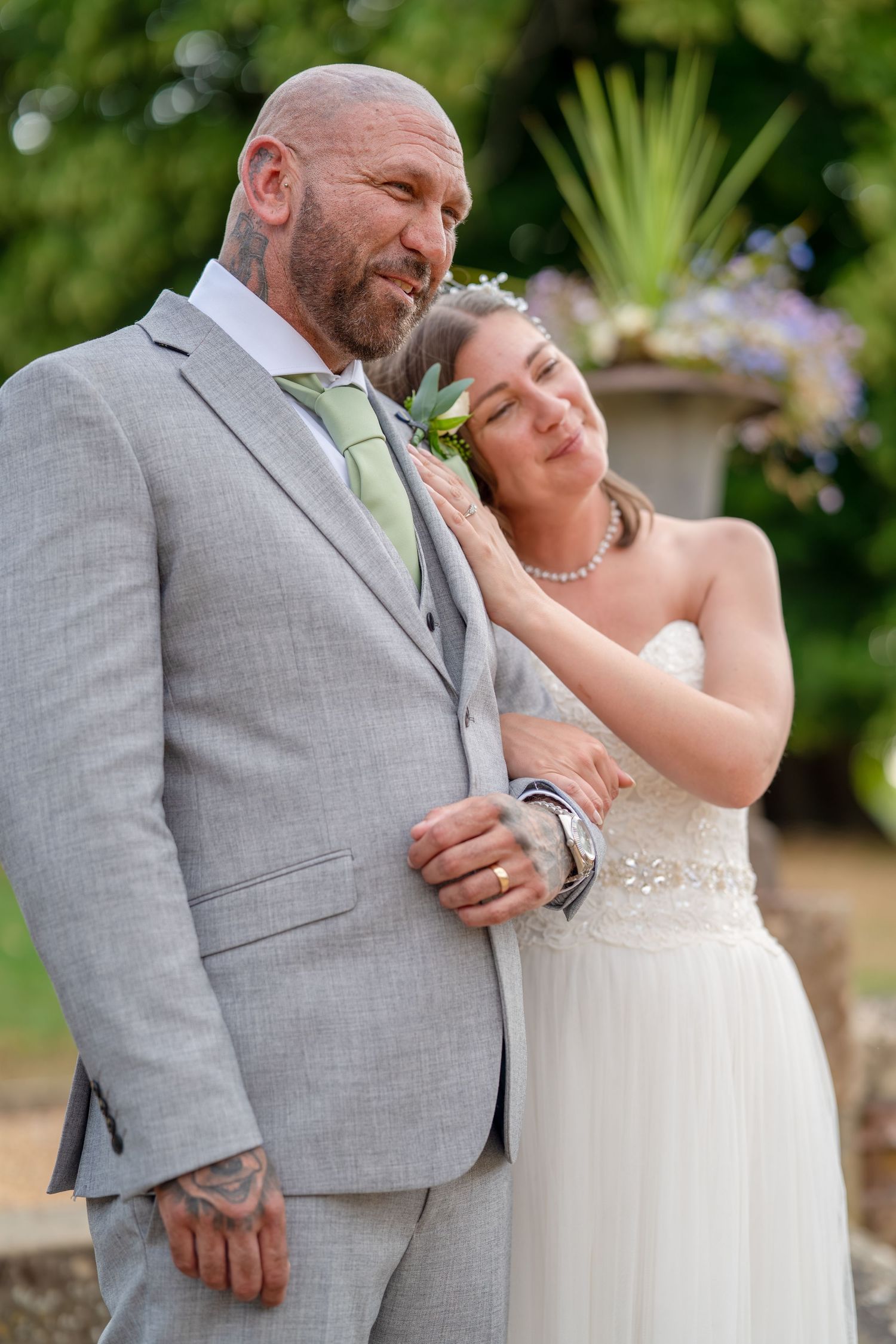 A bride in a white gown rests her hand on a bald groom in a gray suit with a green boutonniere outdoors.