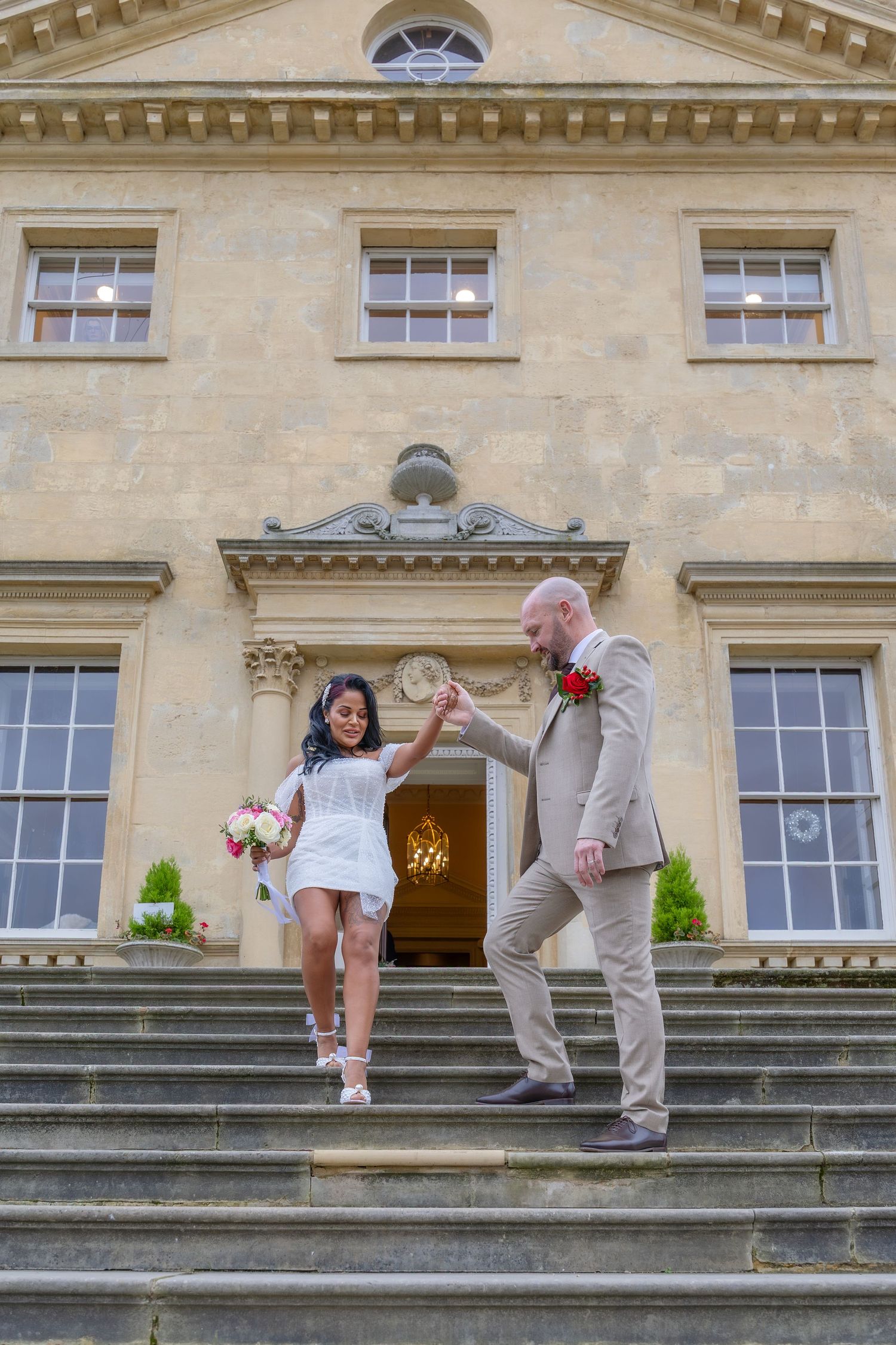 Bride and groom descend grand stone steps of elegant manor house, holding hands as bride lifts leg playfully.