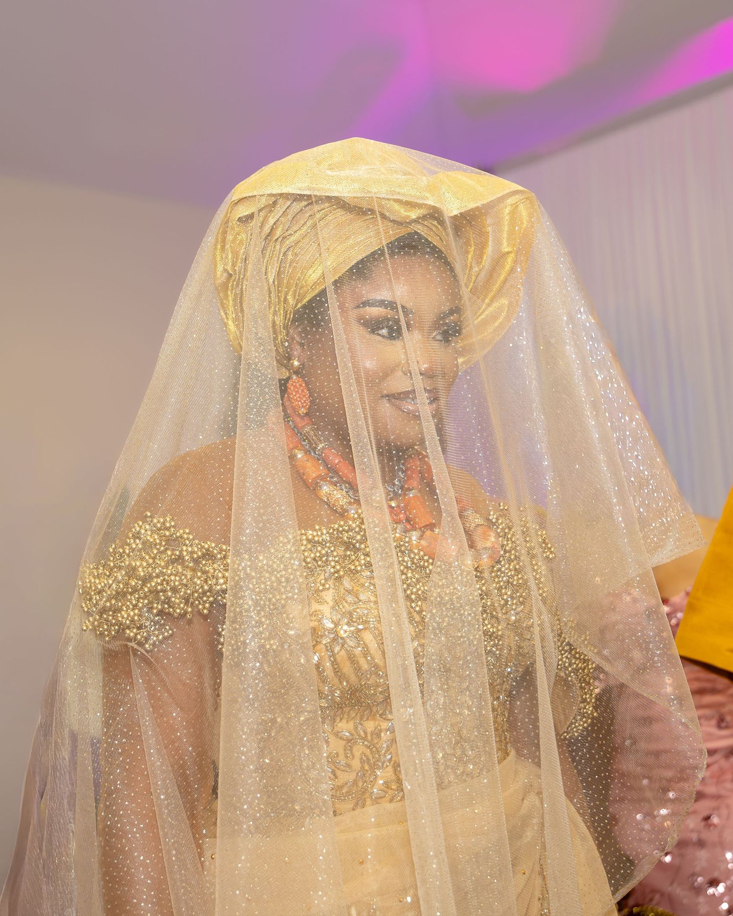 Nigerian bride in gold traditional attire with sparkling veil and coral beaded jewelry at a traditional wedding ceremony.