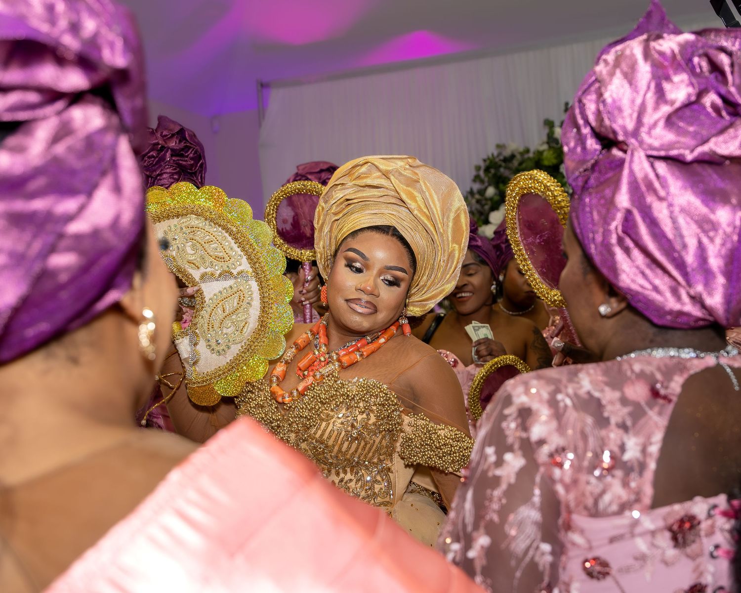Beautiful Nigerian bride in gold gele headwrap and coral beads surrounded by bridesmaids in pink and purple.