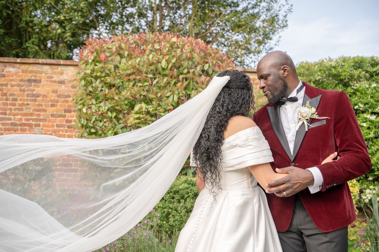 Bride and groom share a romantic kiss outdoors, her veil flowing in the wind, as he wears a red velvet blazer.