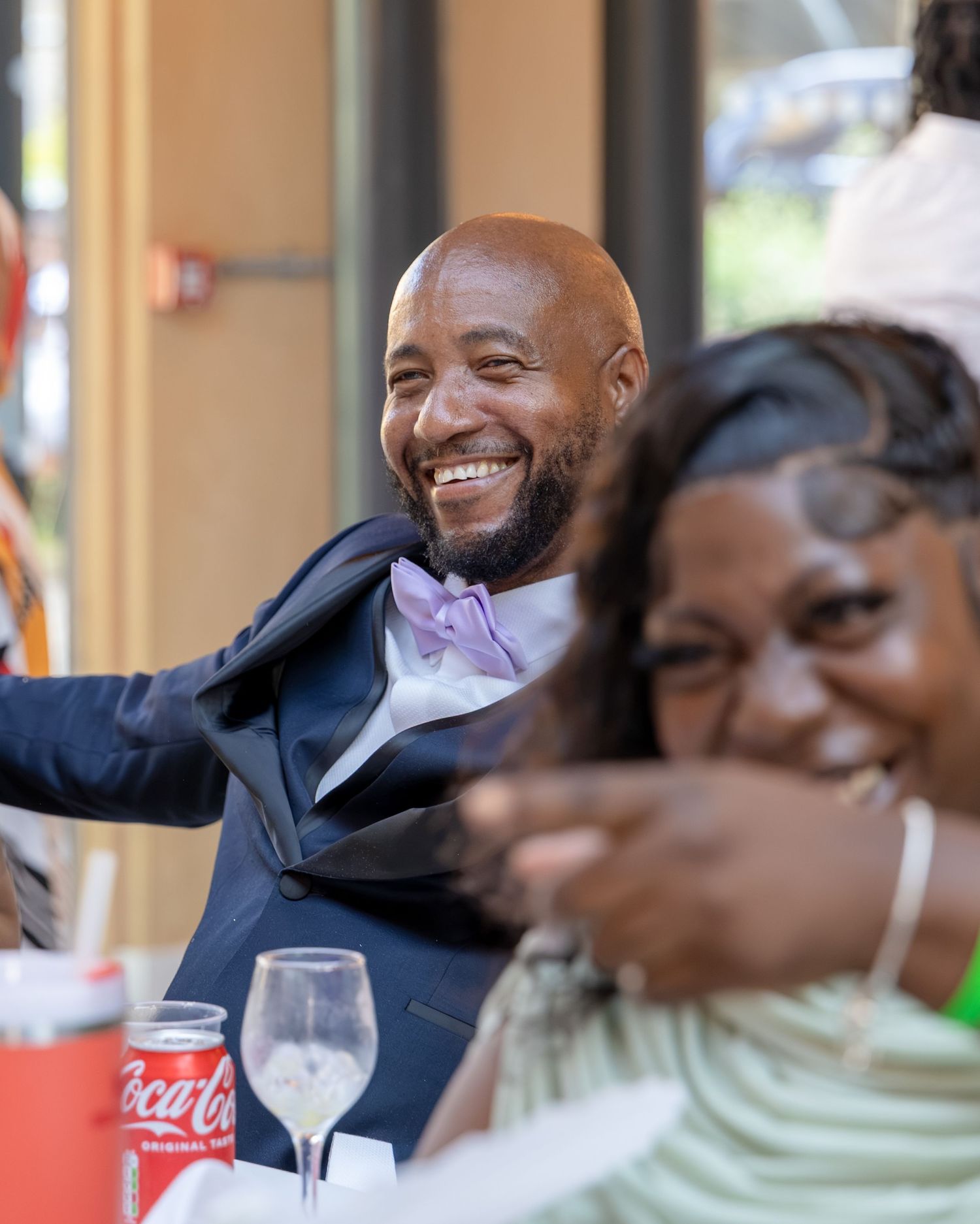 A smiling bald man in a navy suit with purple bow tie laughs joyfully at a formal dinner event.