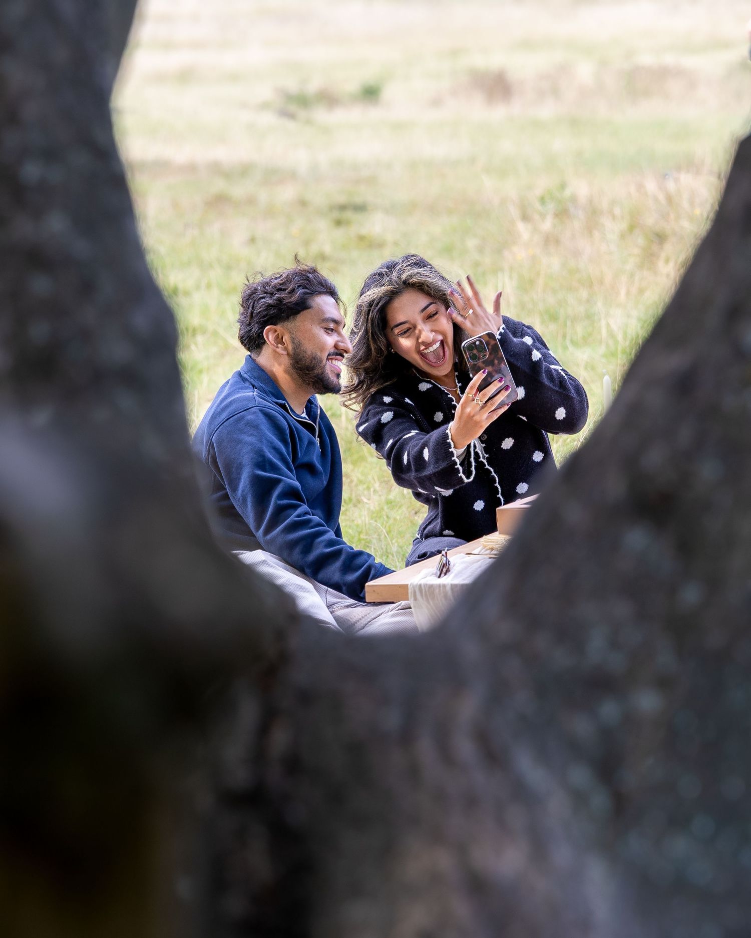 Happy couple celebrating engagement at outdoor picnic, woman displaying ring framed by tree trunks.