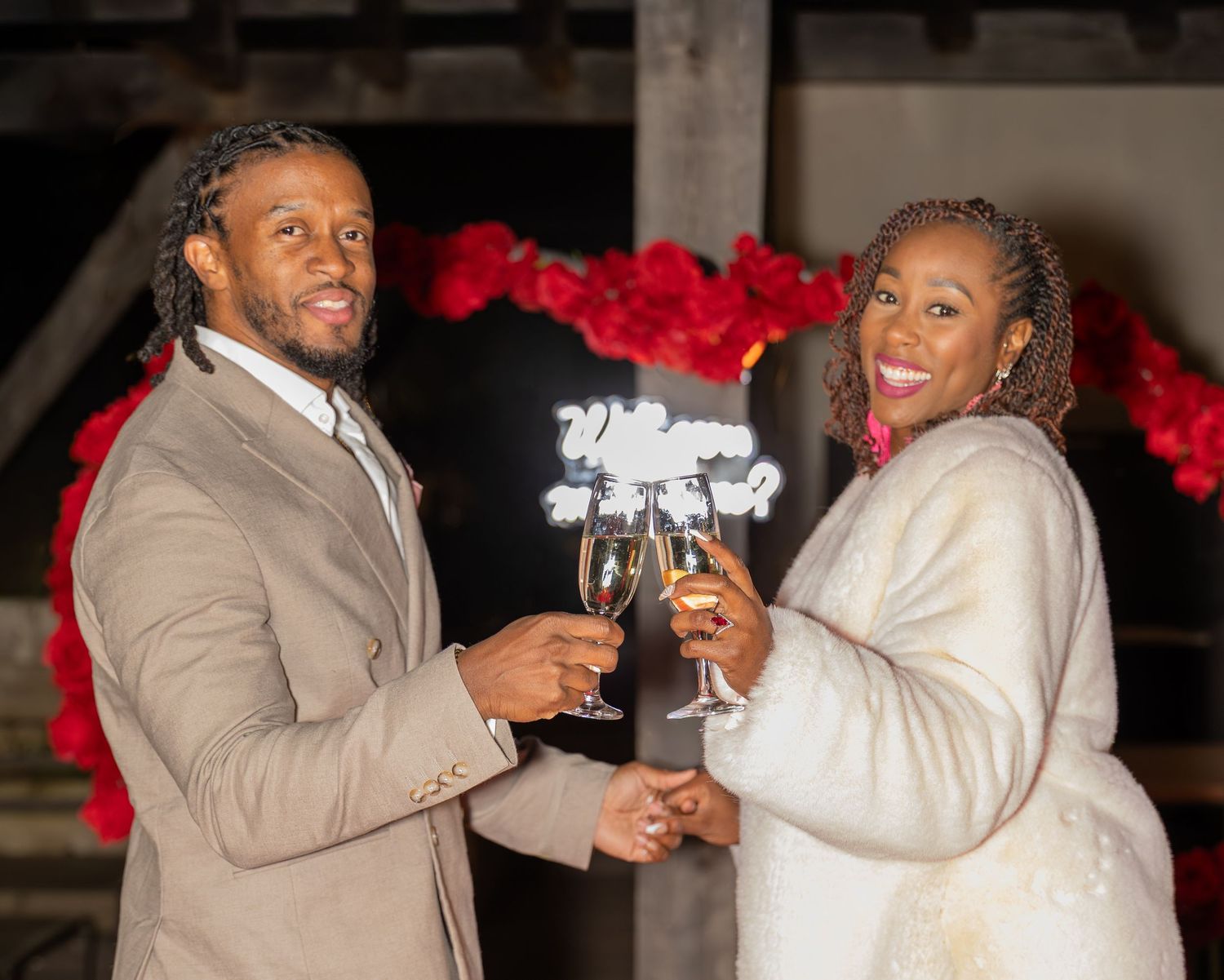 Happy couple toasting with champagne glasses in front of a red floral arch display.