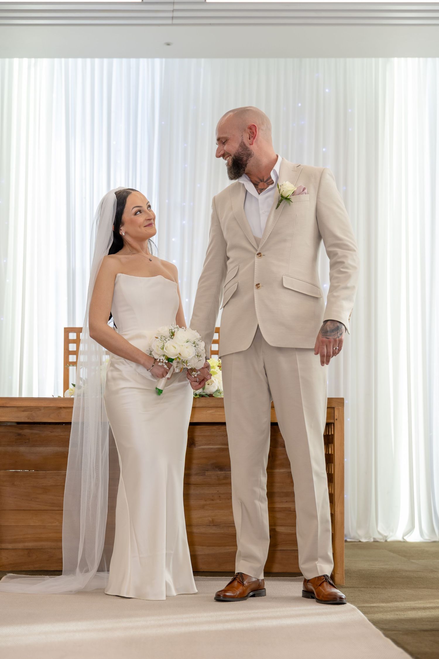 Newlyweds exchange a loving glance at the altar, bride holding white bouquet in elegant satin gown.