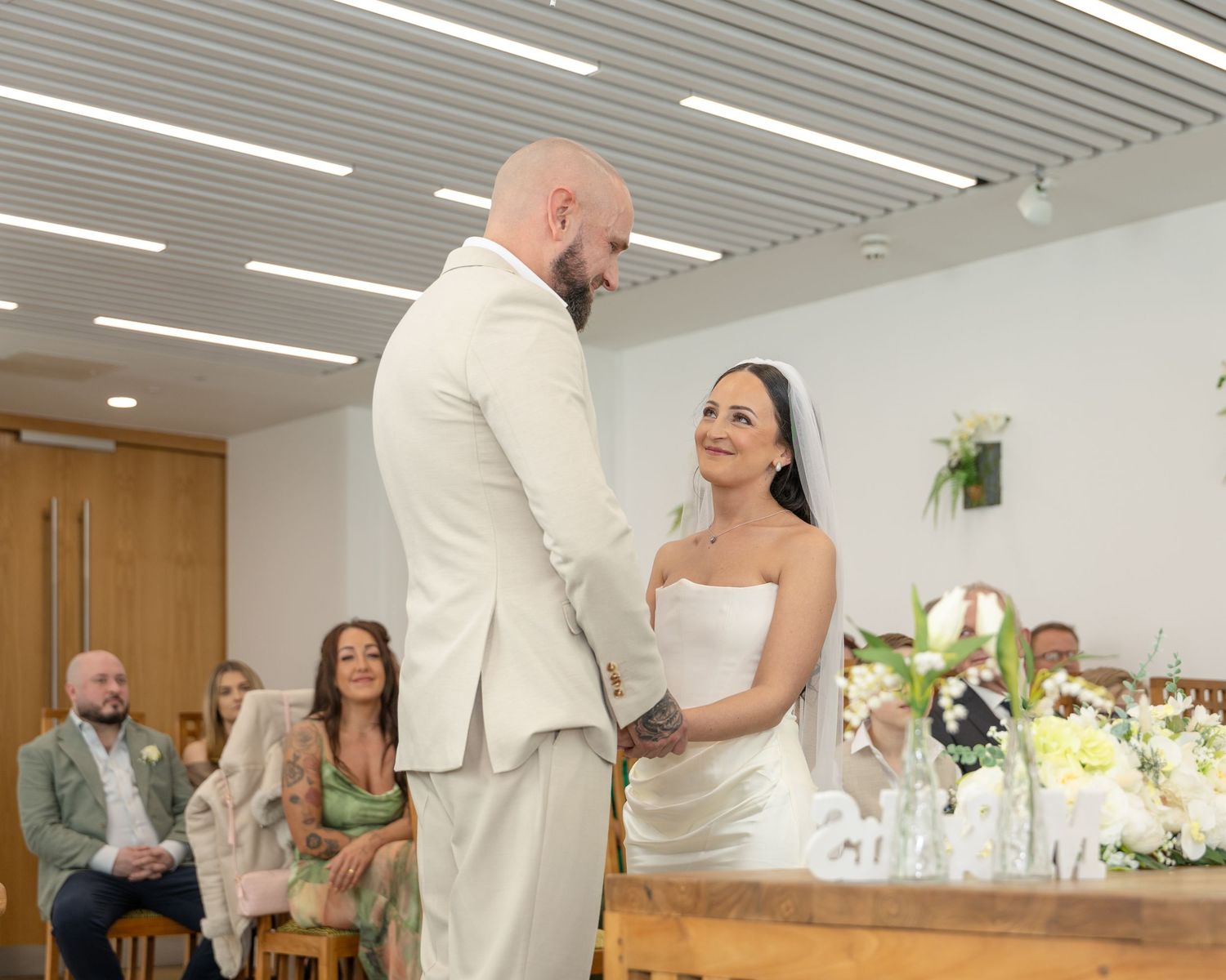 Bald groom in cream suit holds hands with smiling bride in white strapless gown at wedding ceremony altar.