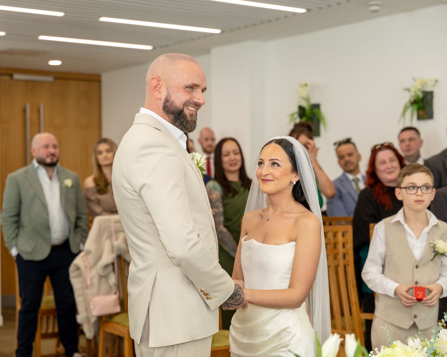 Bald groom in cream suit and veiled bride smile at each other at the altar with guests standing behind.
