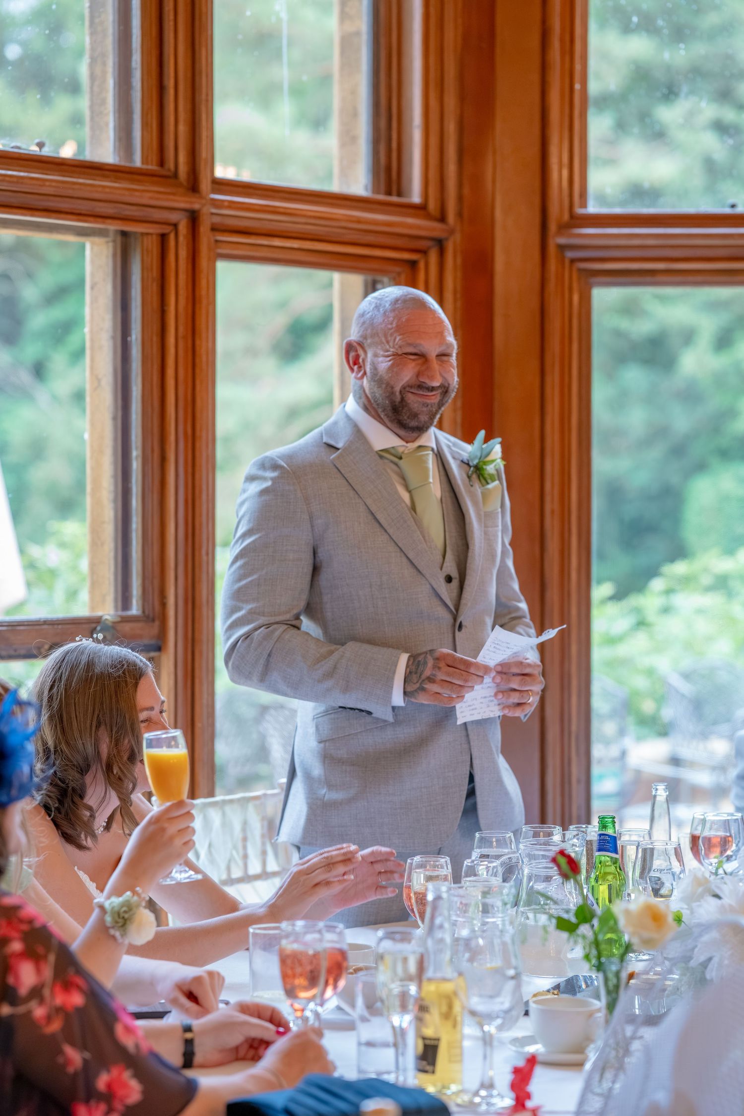 Smiling groom in gray suit giving a speech at wedding reception table with large wooden framed windows behind him.