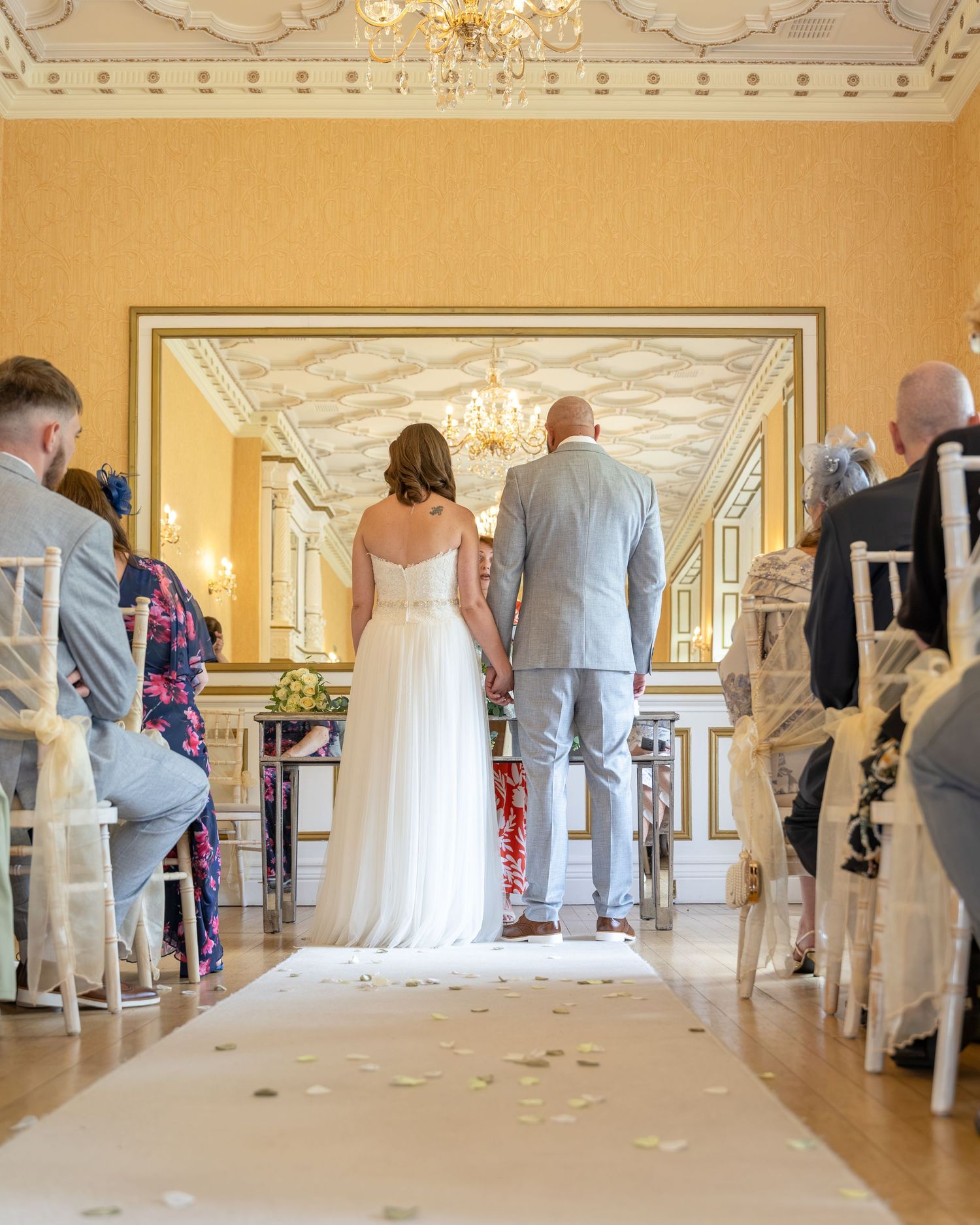 Bride and groom walk down the aisle in an elegant gold wedding venue with chandeliers and white chairs.
