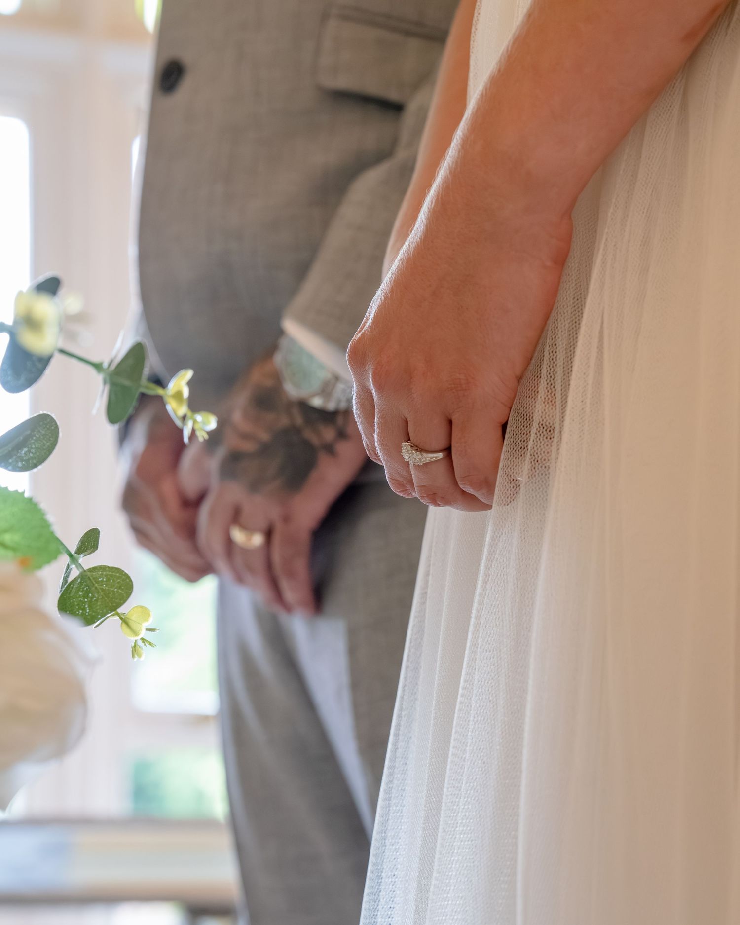 Close-up of a bride and groom holding hands, showing wedding rings, white tulle gown, and gray suit at a wedding ceremony.